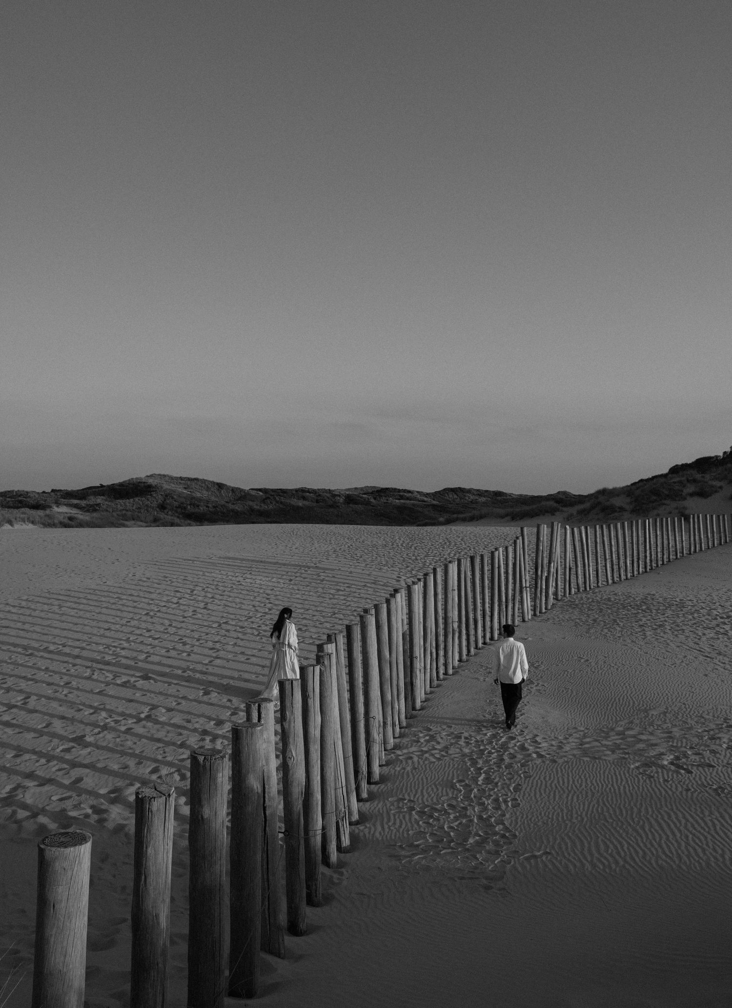 Black and white photo of two people walking along a wooden post fence on a vast sandy beach at dusk.