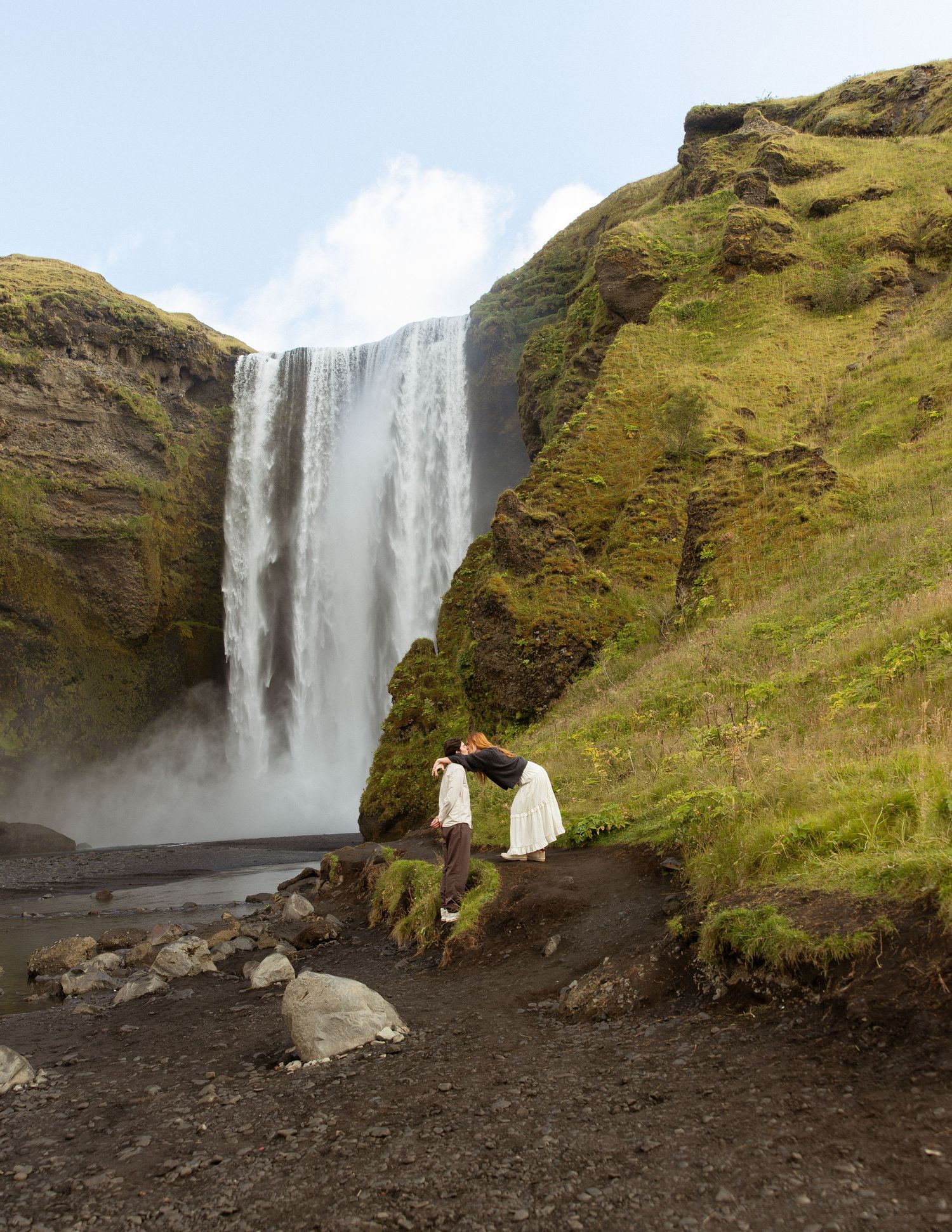 Couple kissing in front of majestic Skogafoss waterfall in Iceland, surrounded by lush green cliffs.