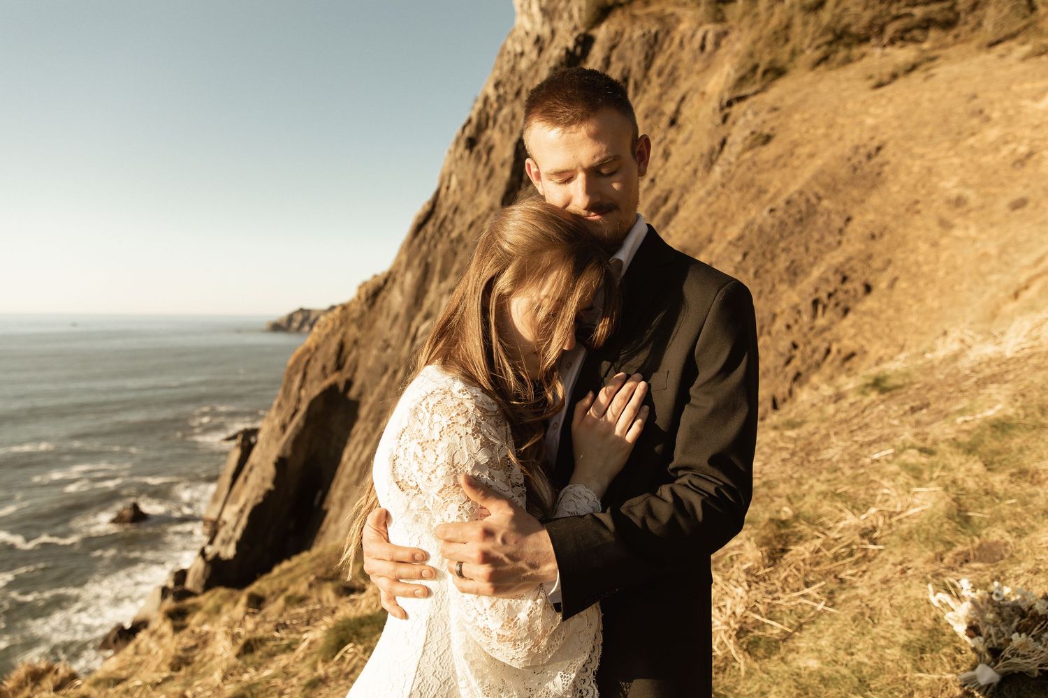 Couple embraces on coastal cliffs overlooking the ocean during golden hour.