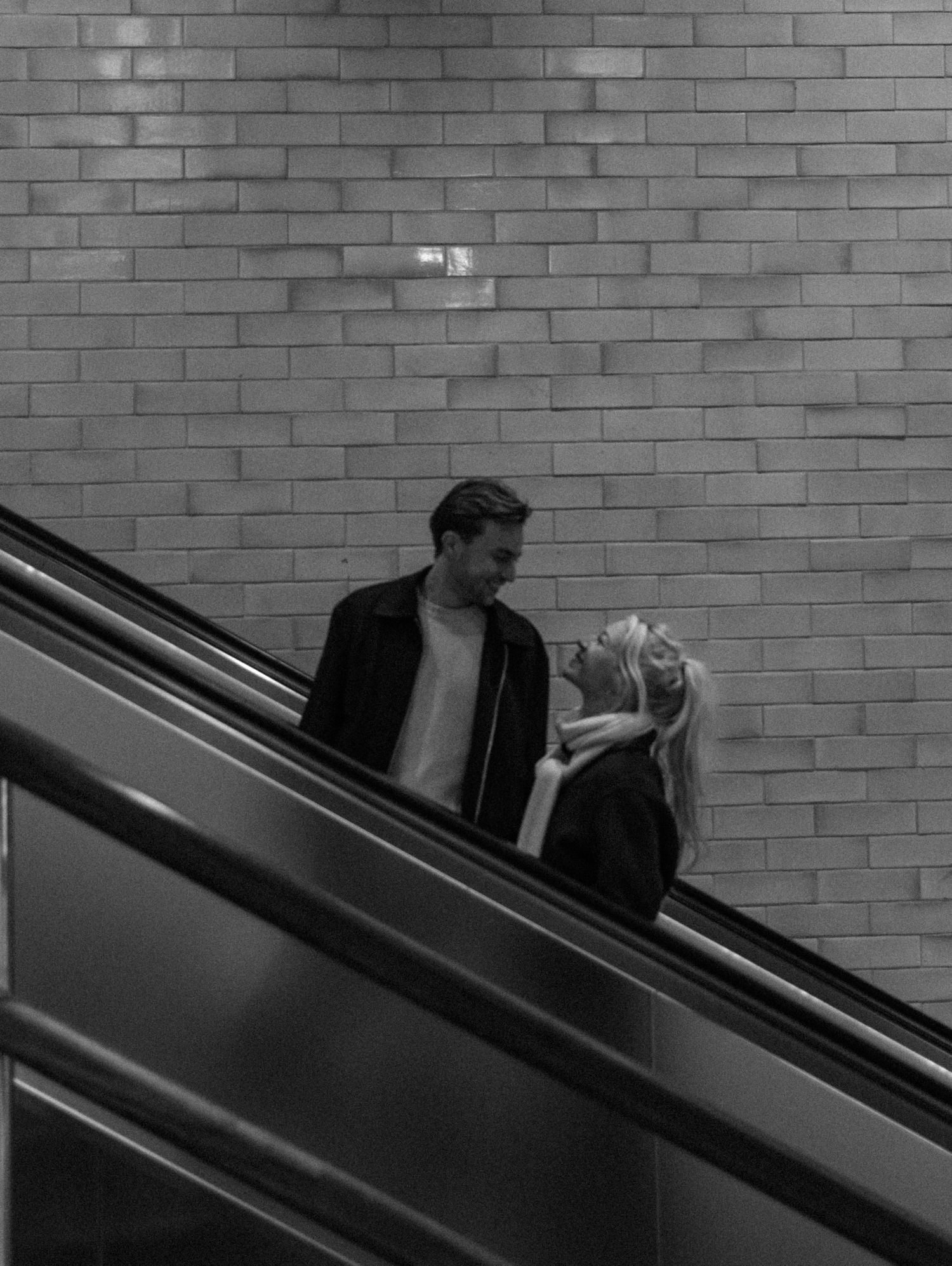 A black and white photo of two people riding an escalator against a brick wall background.