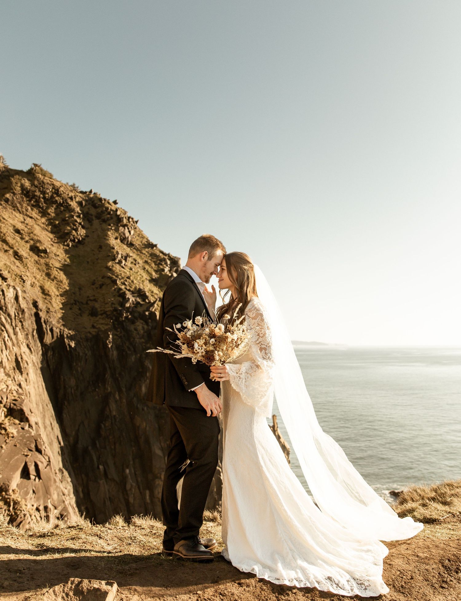 A romantic wedding couple embraces on a scenic coastal cliff overlooking the ocean at sunset.