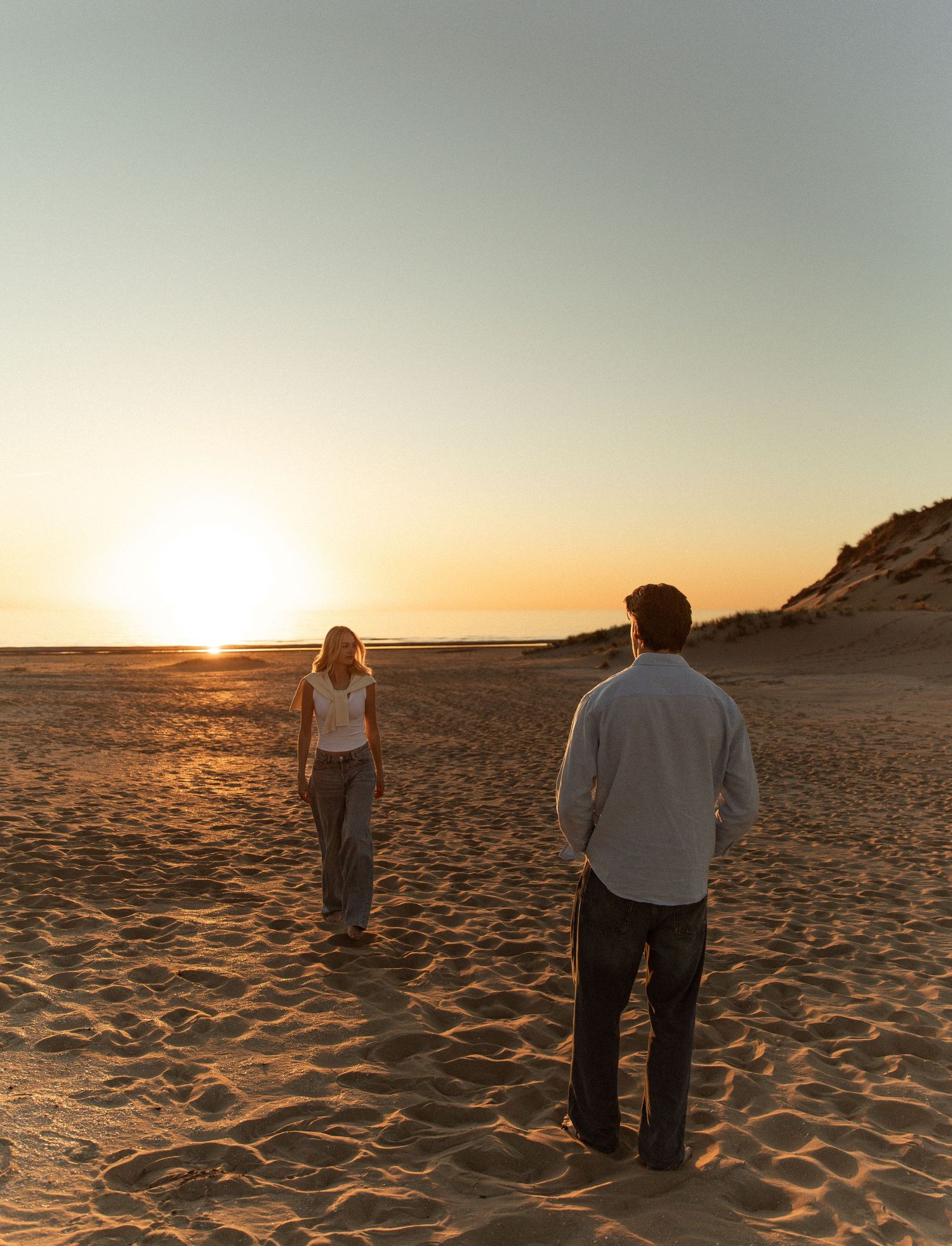A couple walks on a sandy beach at sunset, with golden light reflecting off the sand and calm ocean horizon.