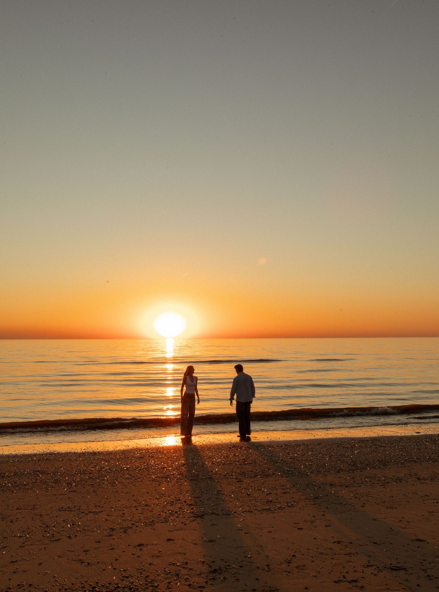 Couple silhouetted against a golden sunset on a tranquil beach, with the sun reflecting on calm ocean waters.