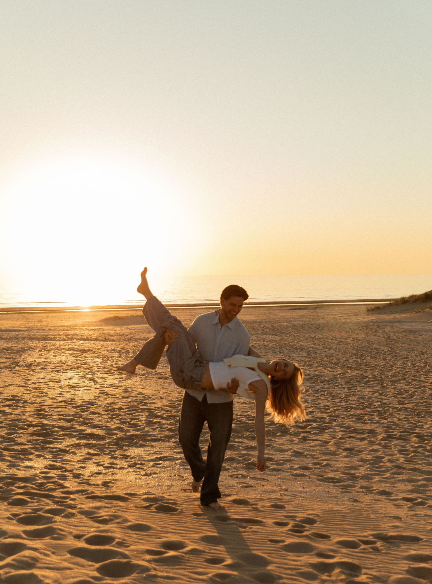Couple dancing playfully on a sandy beach at golden sunset, man lifting woman in romantic outdoor photo.