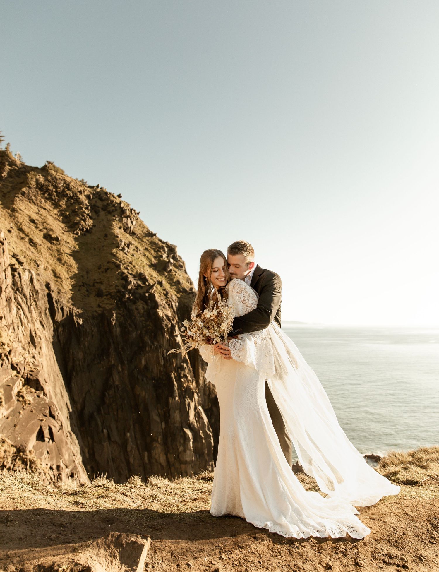 Couple in wedding attire embracing on rocky coastal cliffs at sunset with ocean views in the background.
