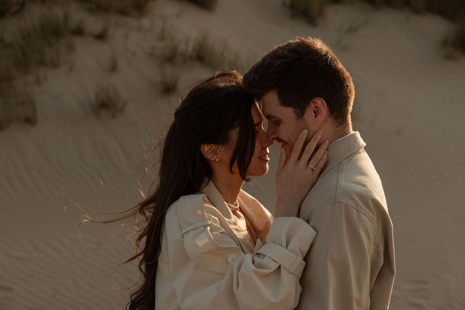 Romantic couple embracing in sandy dunes at golden hour, foreheads touching tenderly in warm desert light.