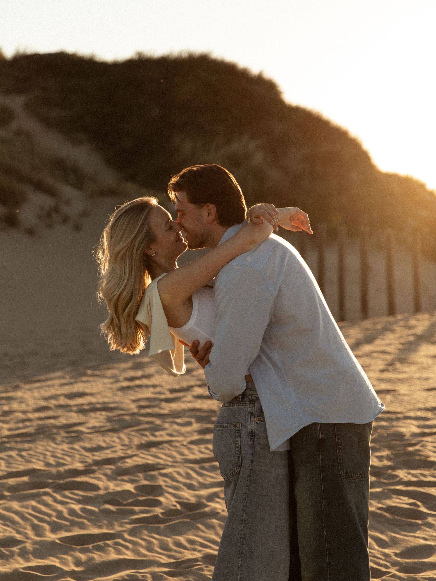 Couple embracing and kissing on a sandy beach at golden sunset with sand dunes in the background.