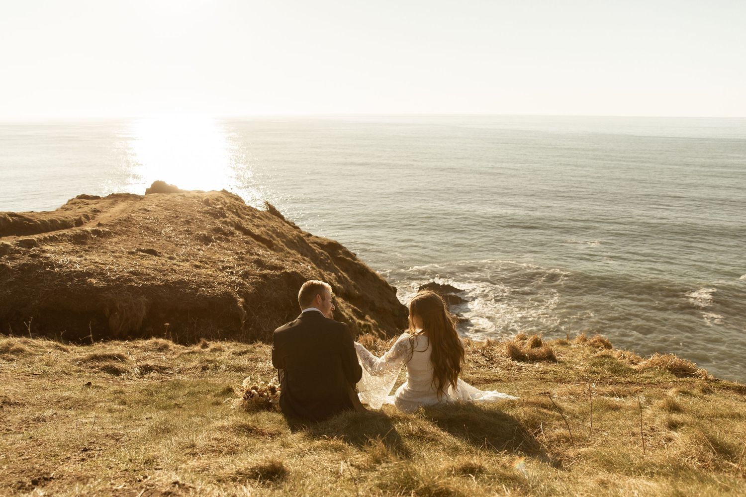 A person sits on a coastal cliff overlooking a misty ocean view at sunset.