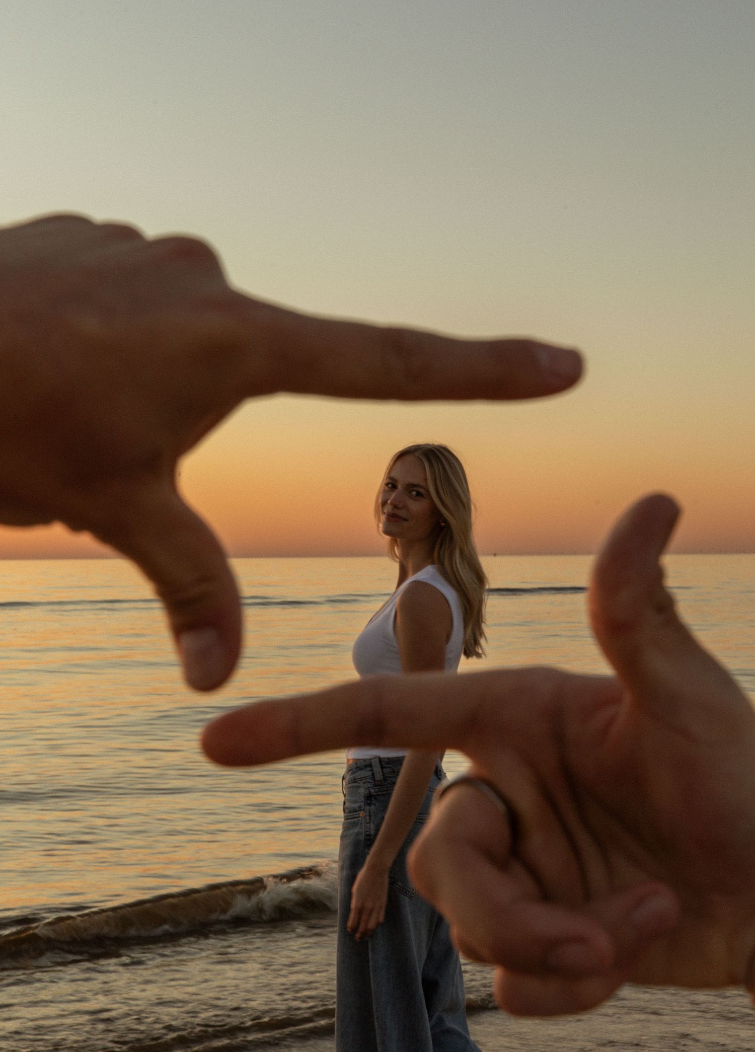 Woman standing on beach at sunset framed by hands forming a camera viewfinder gesture.