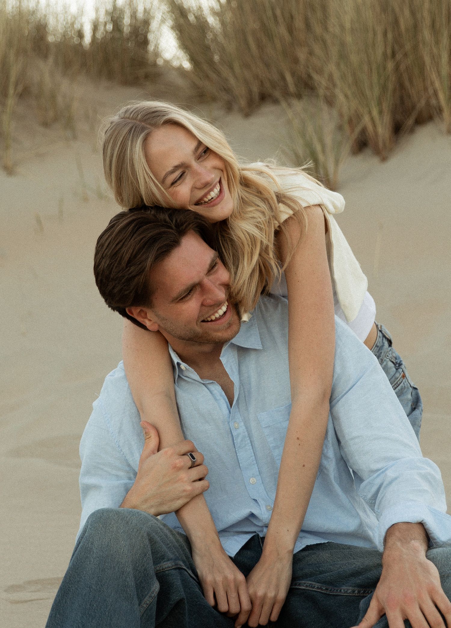 Happy couple laughing and embracing on sandy beach dunes, woman with blonde hair hugging man from behind.