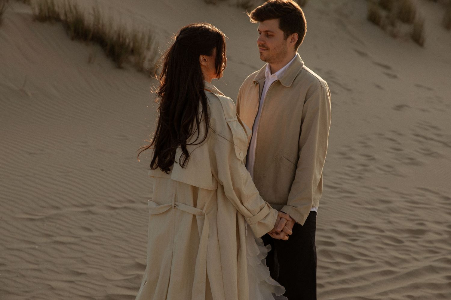 Couple holding hands facing each other on sandy dunes, woman in white trench coat, man in beige jacket, golden hour light.