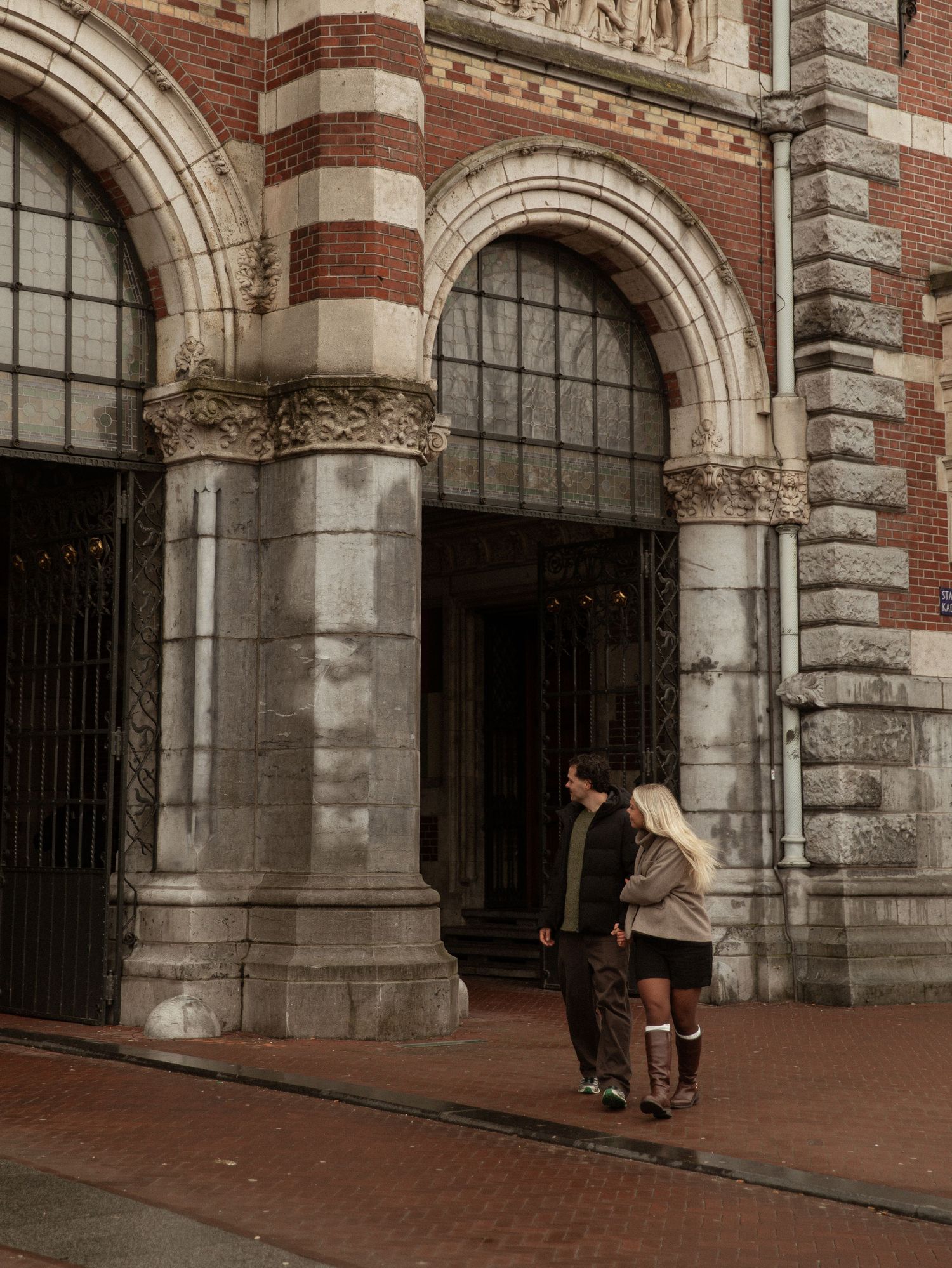 People walk past a historic brick building with arched stone doorways and ornate architectural details.