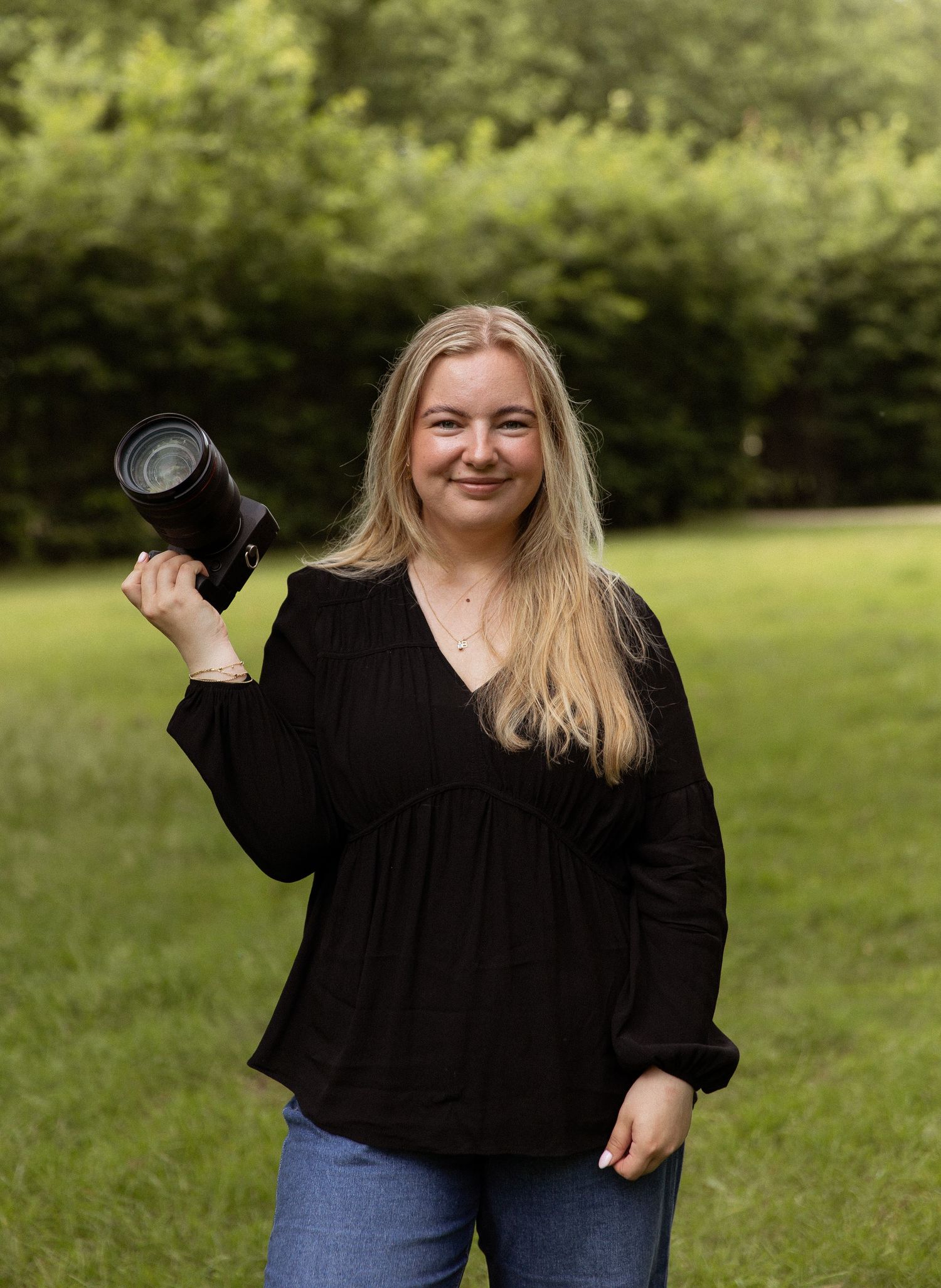 Smiling blonde woman holding a camera lens outdoors in a green park, wearing a black top and jeans.