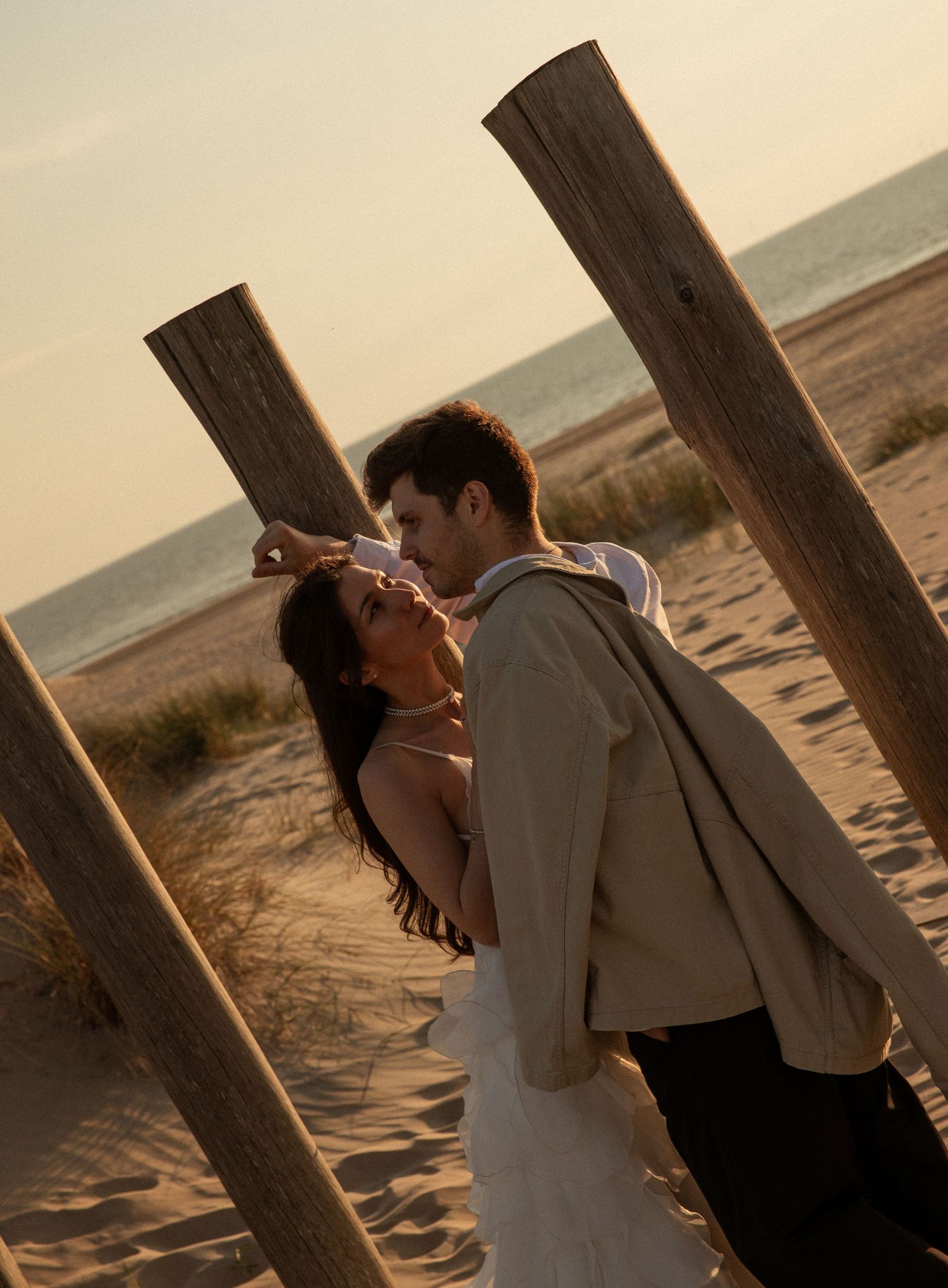 Couple sharing a romantic moment between wooden posts on a sandy beach at golden sunset hour.