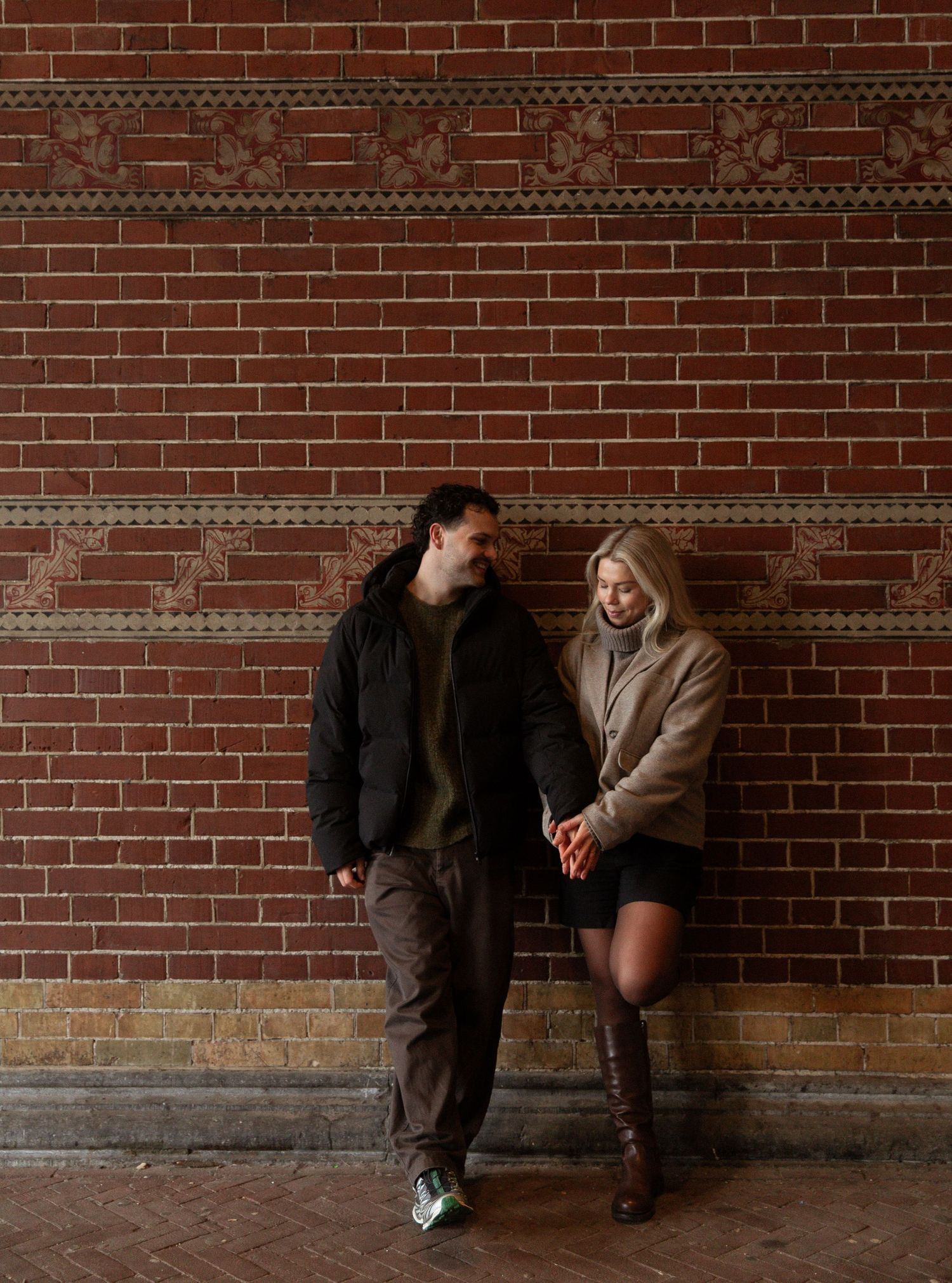 A couple sits intimately against a rustic brick wall in a romantic engagement photo series.