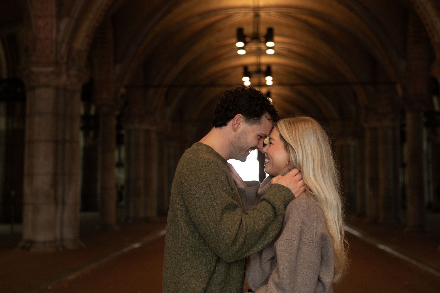 A couple shares a romantic kiss under the arches of a dimly lit stone passageway.