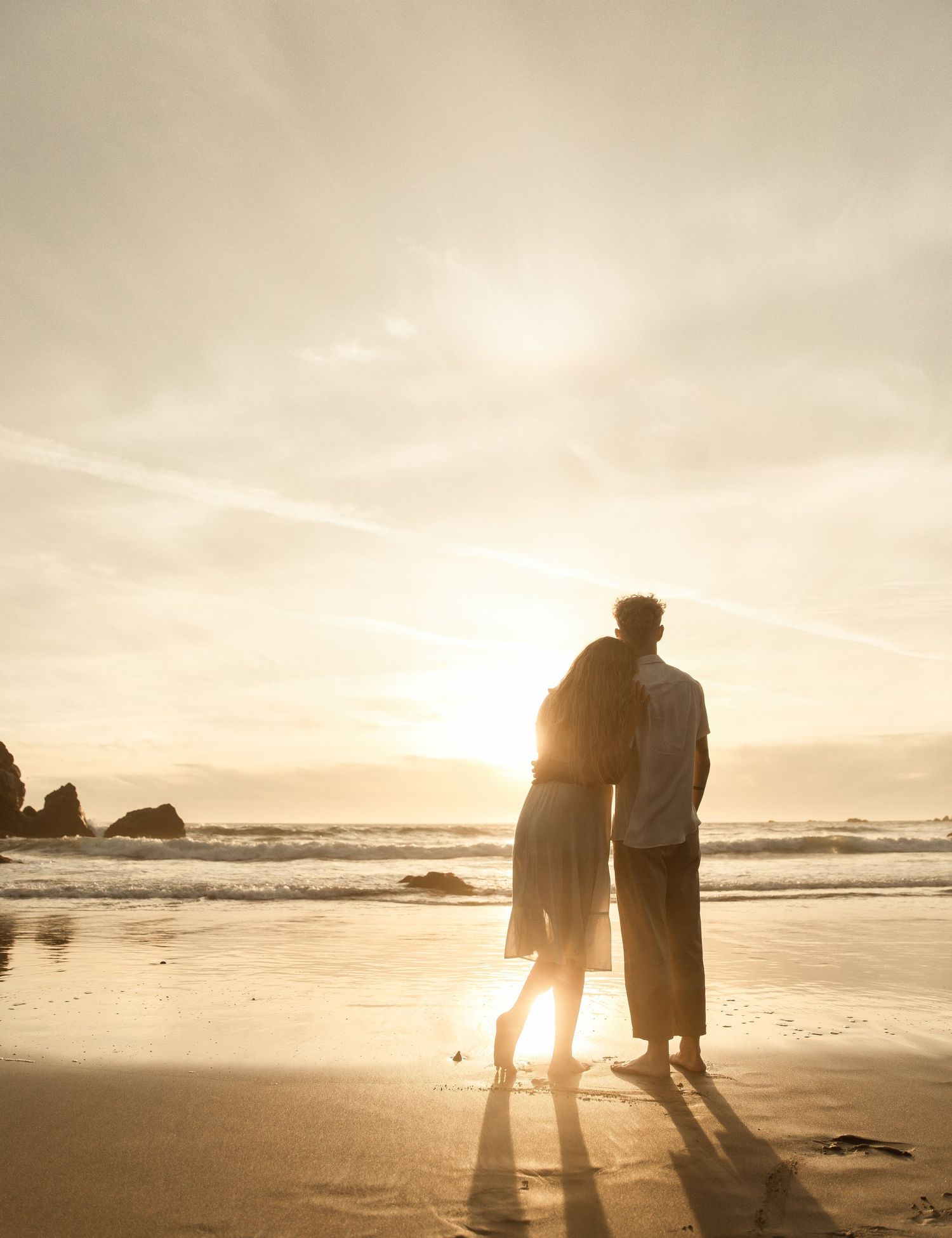 A couple walking along a misty beach at sunset casting long shadows on the sand as waves roll in near the shore.