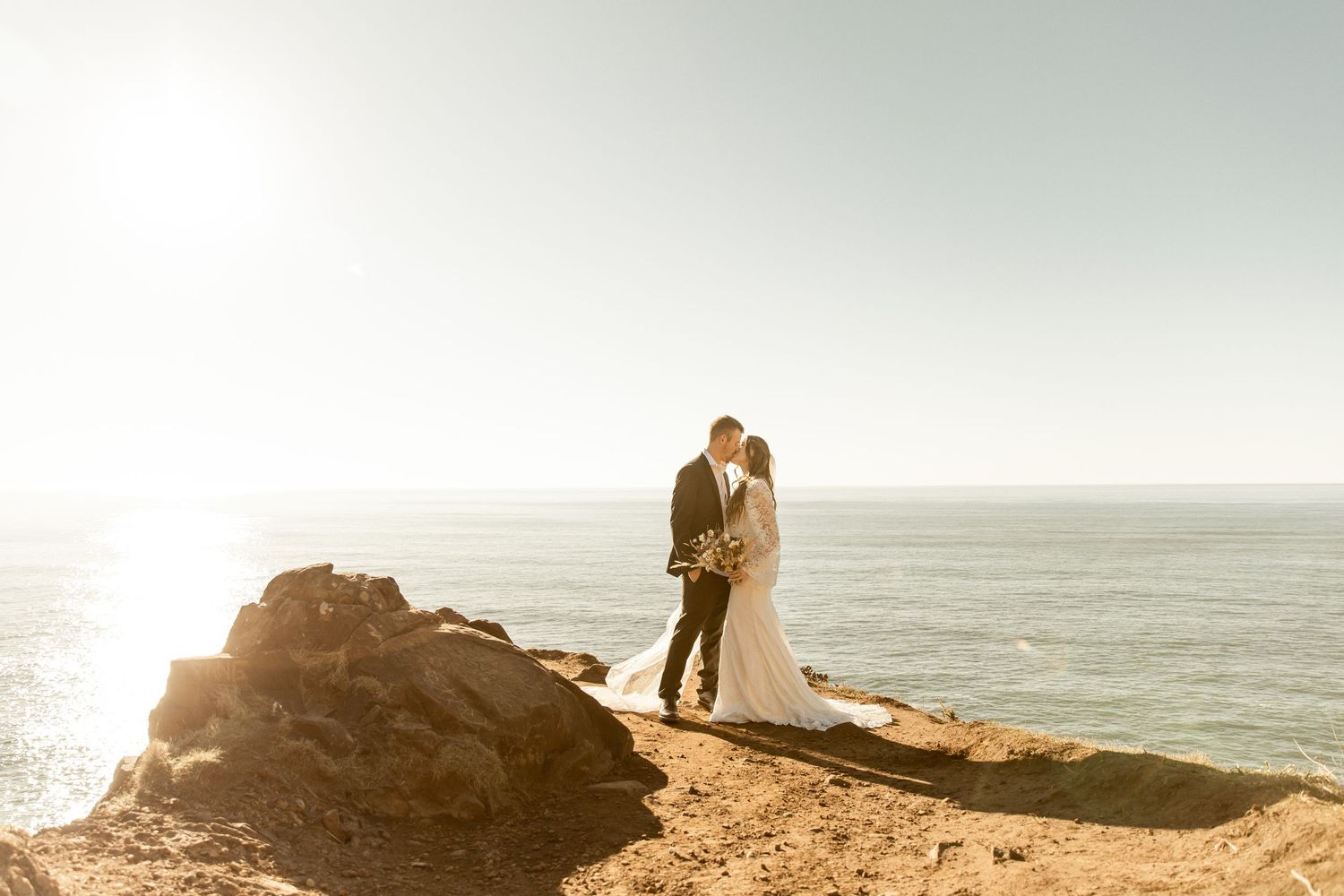 A couple shares a romantic embrace on a coastal cliff overlooking the ocean during golden hour.