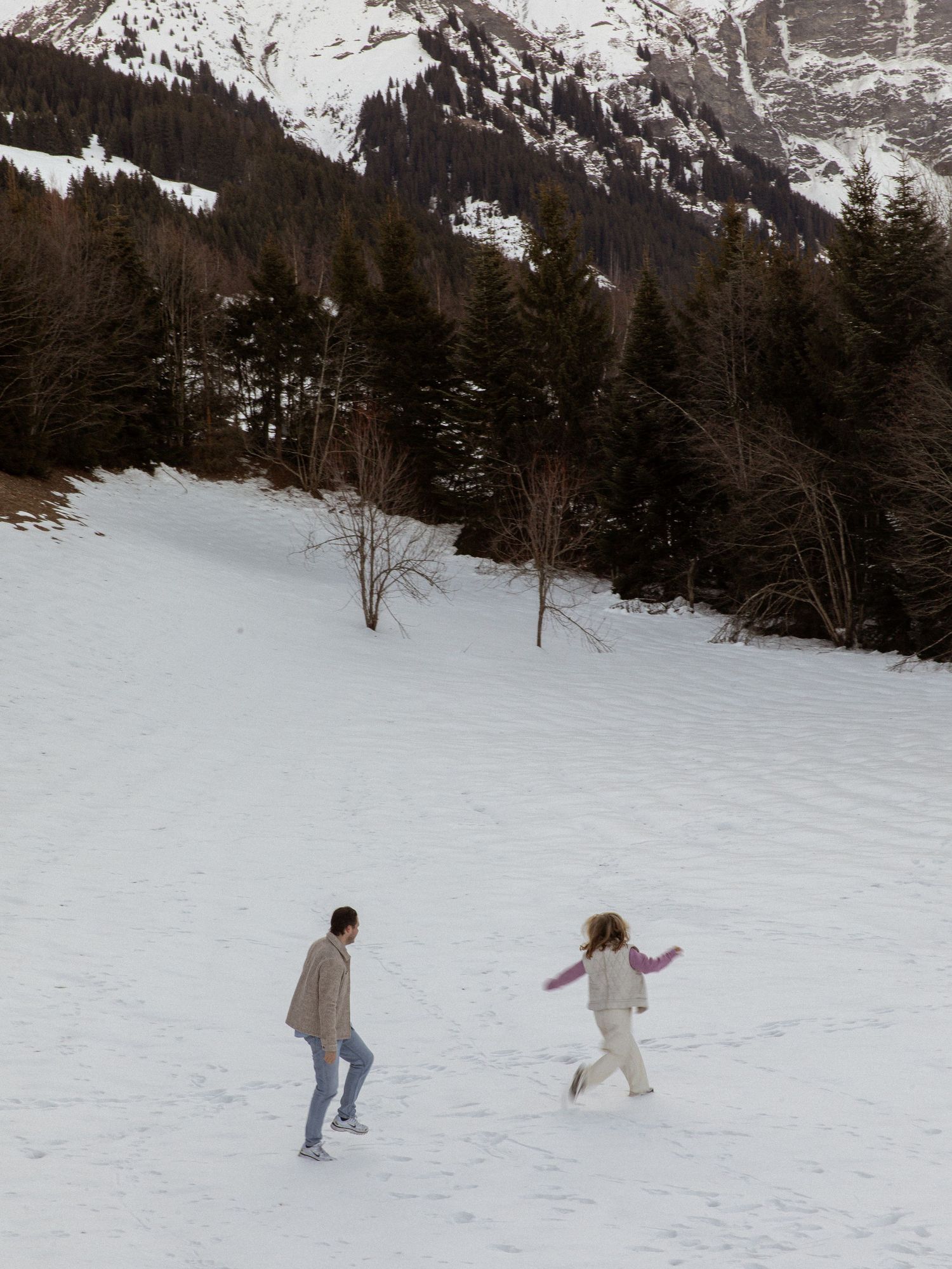 Two people playfully walking through snow with mountains in the background.