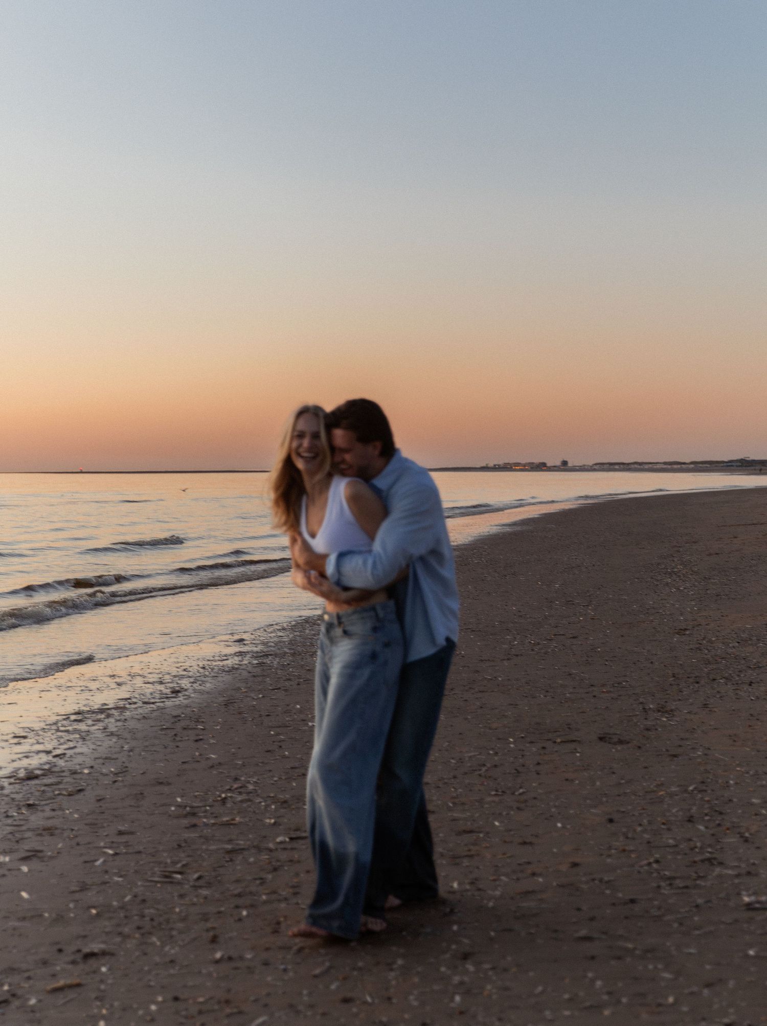 Couple embracing on a sandy beach at sunset with soft pink and orange hues reflecting over calm ocean waters.
