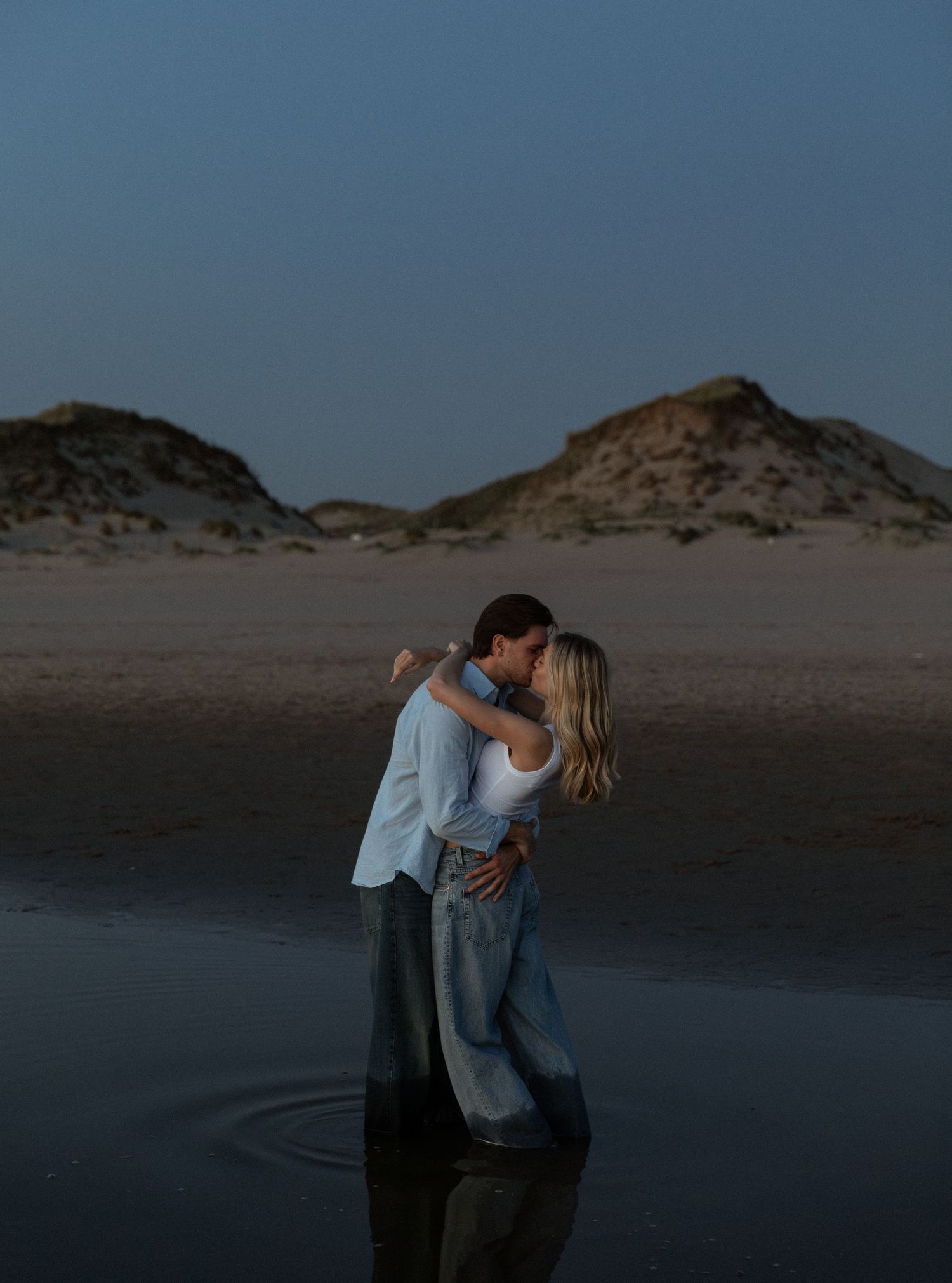 A couple shares a romantic kiss while standing in shallow water on a beach at dusk with sand dunes in the background.