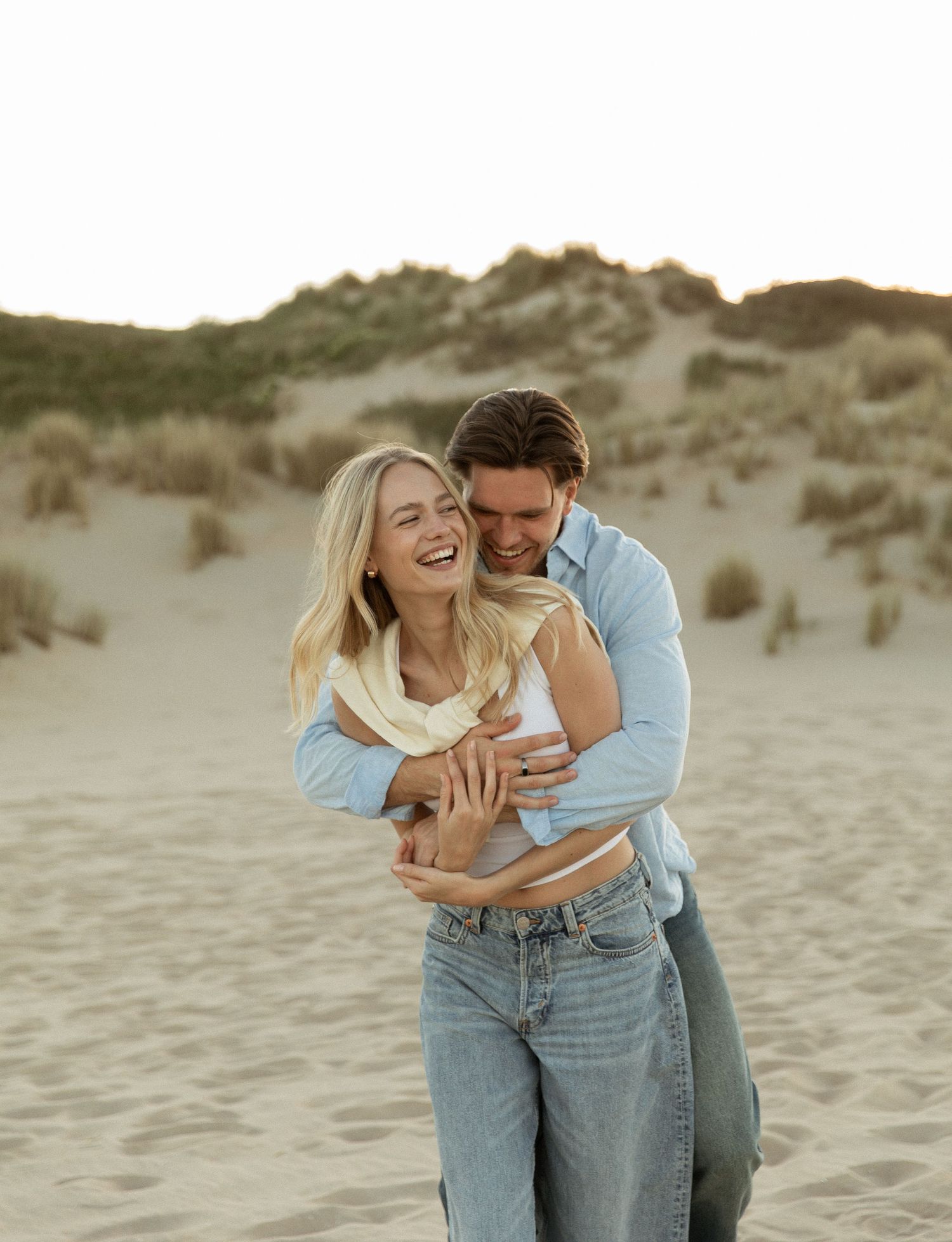 Happy couple embracing and laughing on sandy beach dunes at golden hour, wearing casual blue outfits.