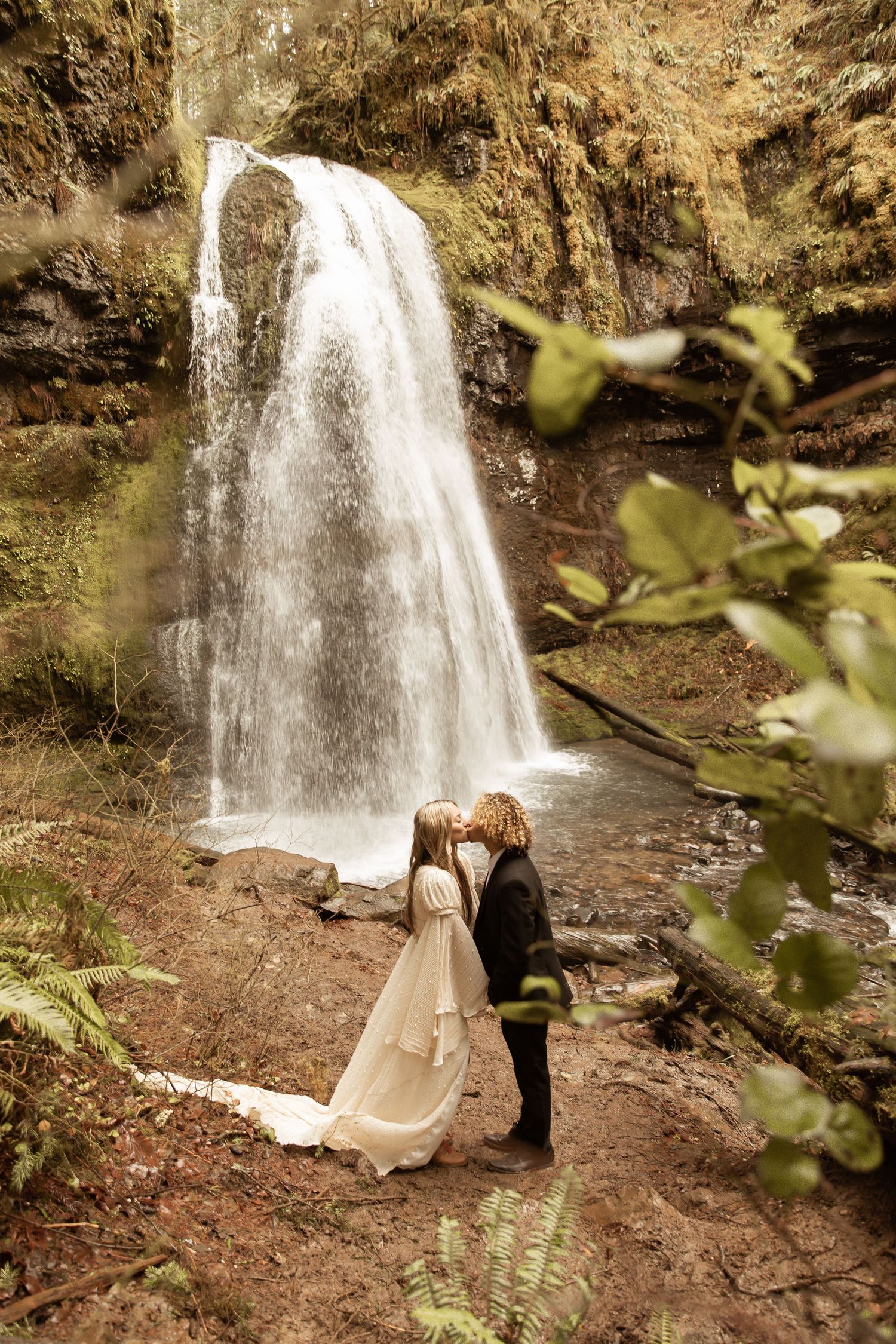 Intimate moment captured by waterfall with flowing white dress against natural scenery.