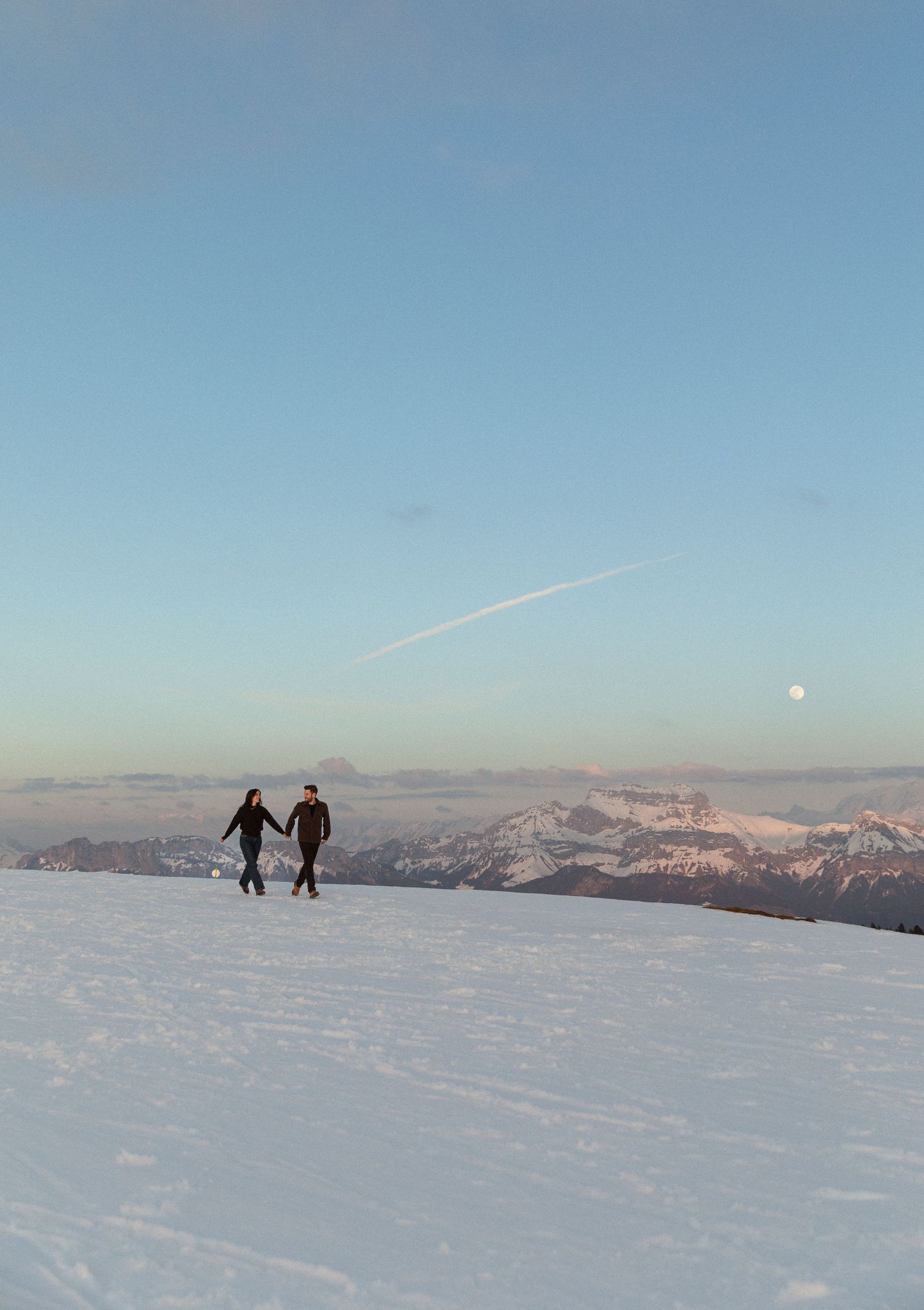 Silhouettes of people walking on a snowy mountain ridge with distant peaks and blue sky in the background.