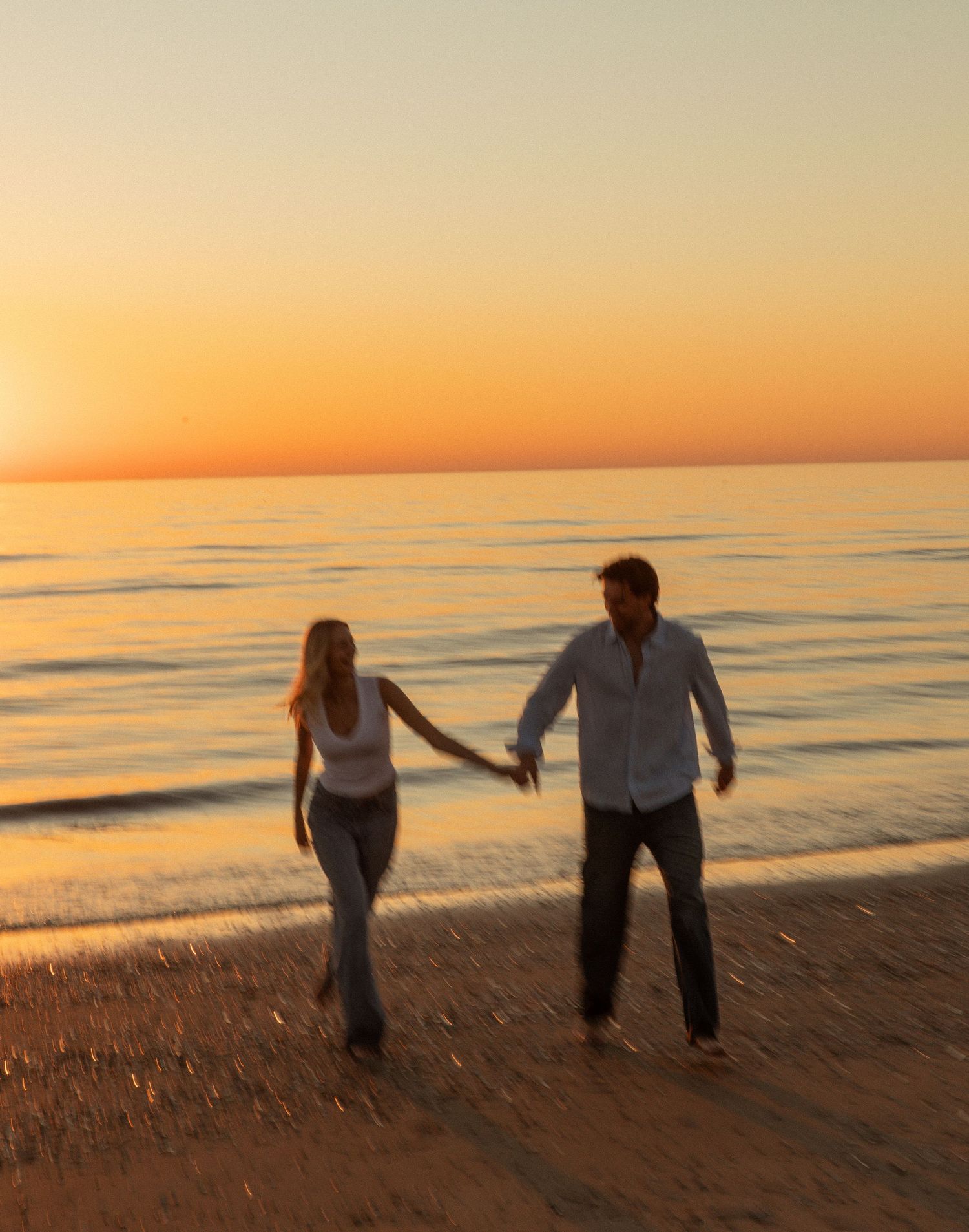 Couple holding hands and walking on a sandy beach at golden sunset with calm ocean waves in the background.