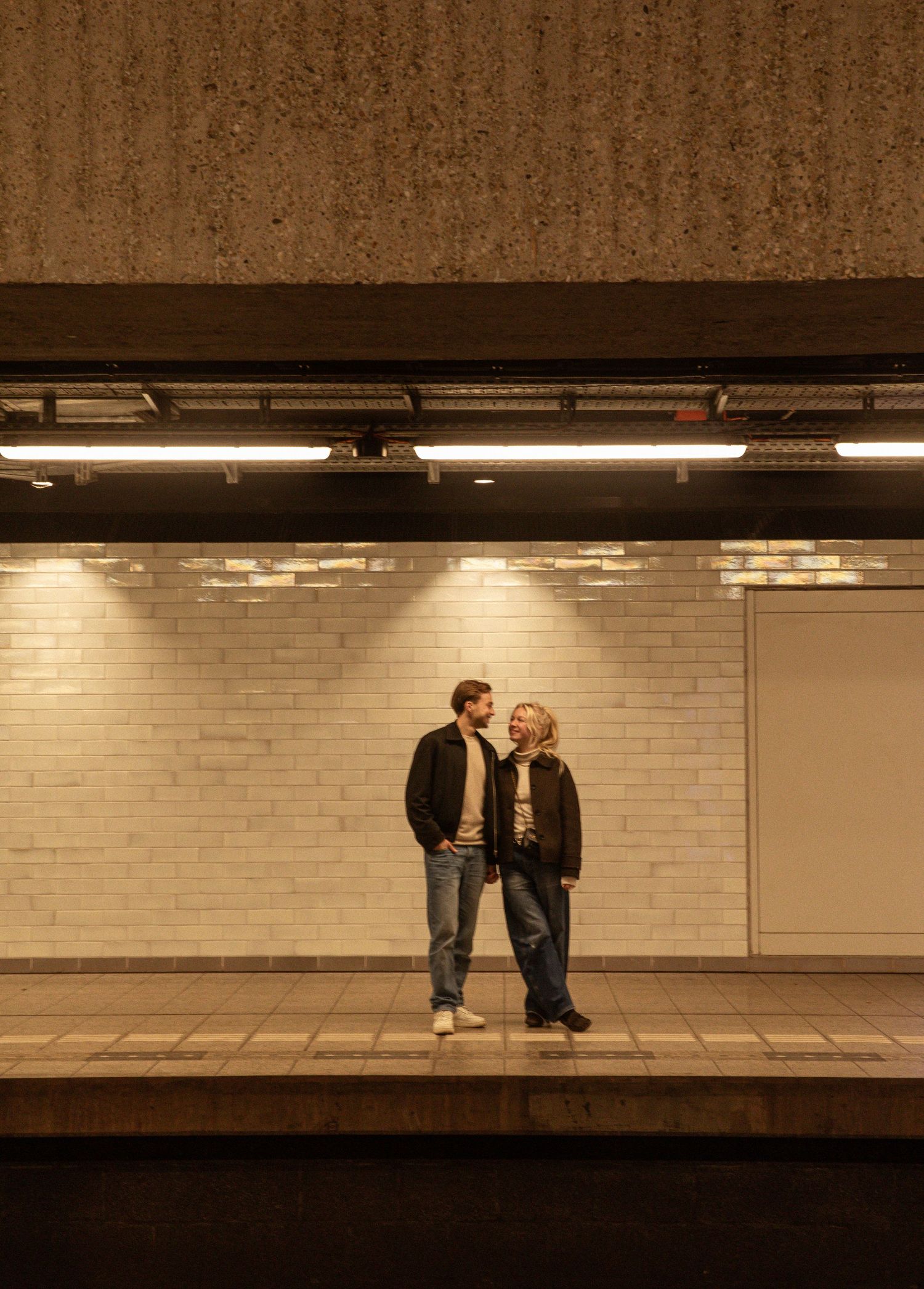 Two people walk past a white brick wall in a subway station with fluorescent lighting overhead.