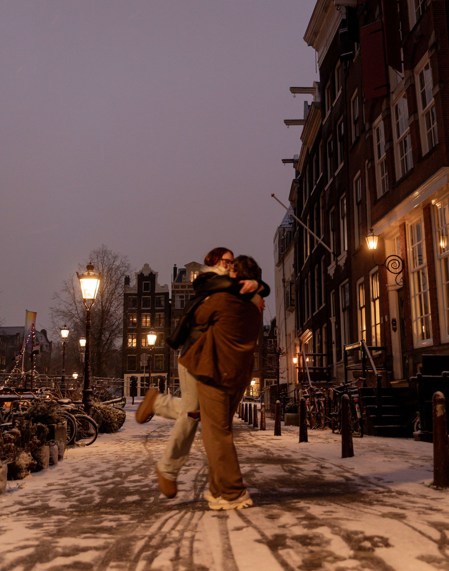 Snowy evening in Amsterdam with lit street lamps and historic buildings.