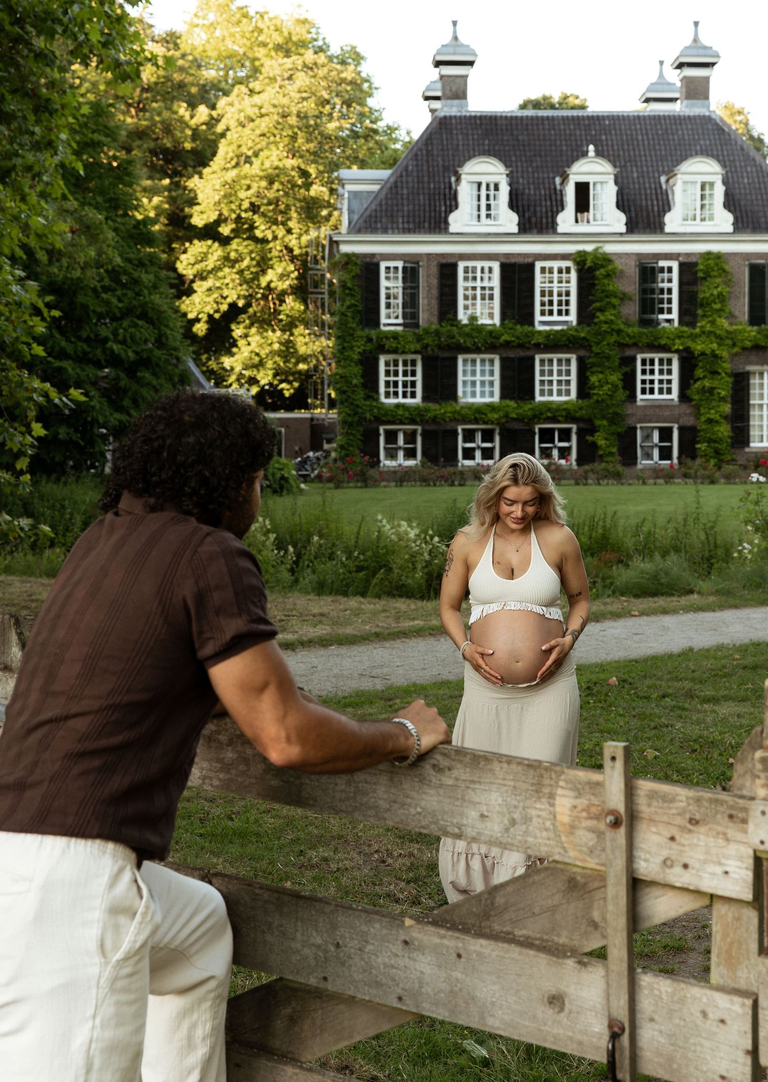 Pregnant woman in white crop top holds baby bump near wooden fence, ivy-covered brick manor house in background.