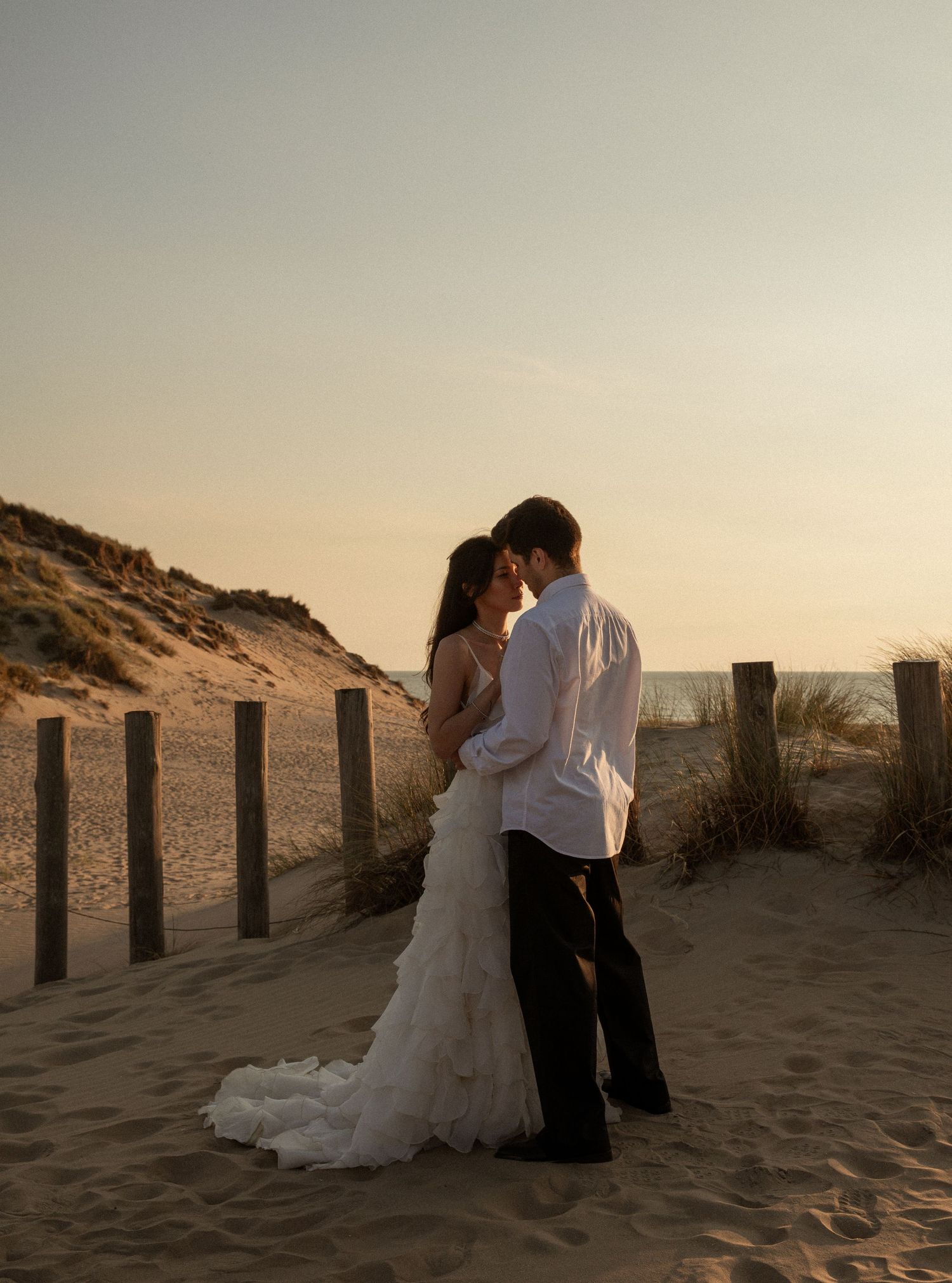 Bride and groom embrace on sandy beach path at sunset, wedding dress trailing on sand with ocean in background.