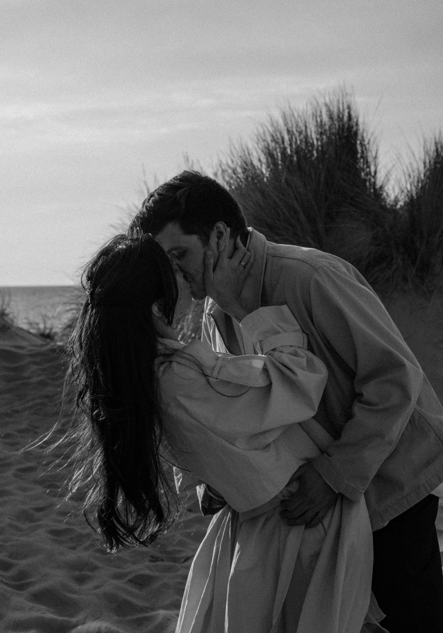 Black and white photo of a couple sharing a romantic kiss on a sandy beach with grass dunes in the background.