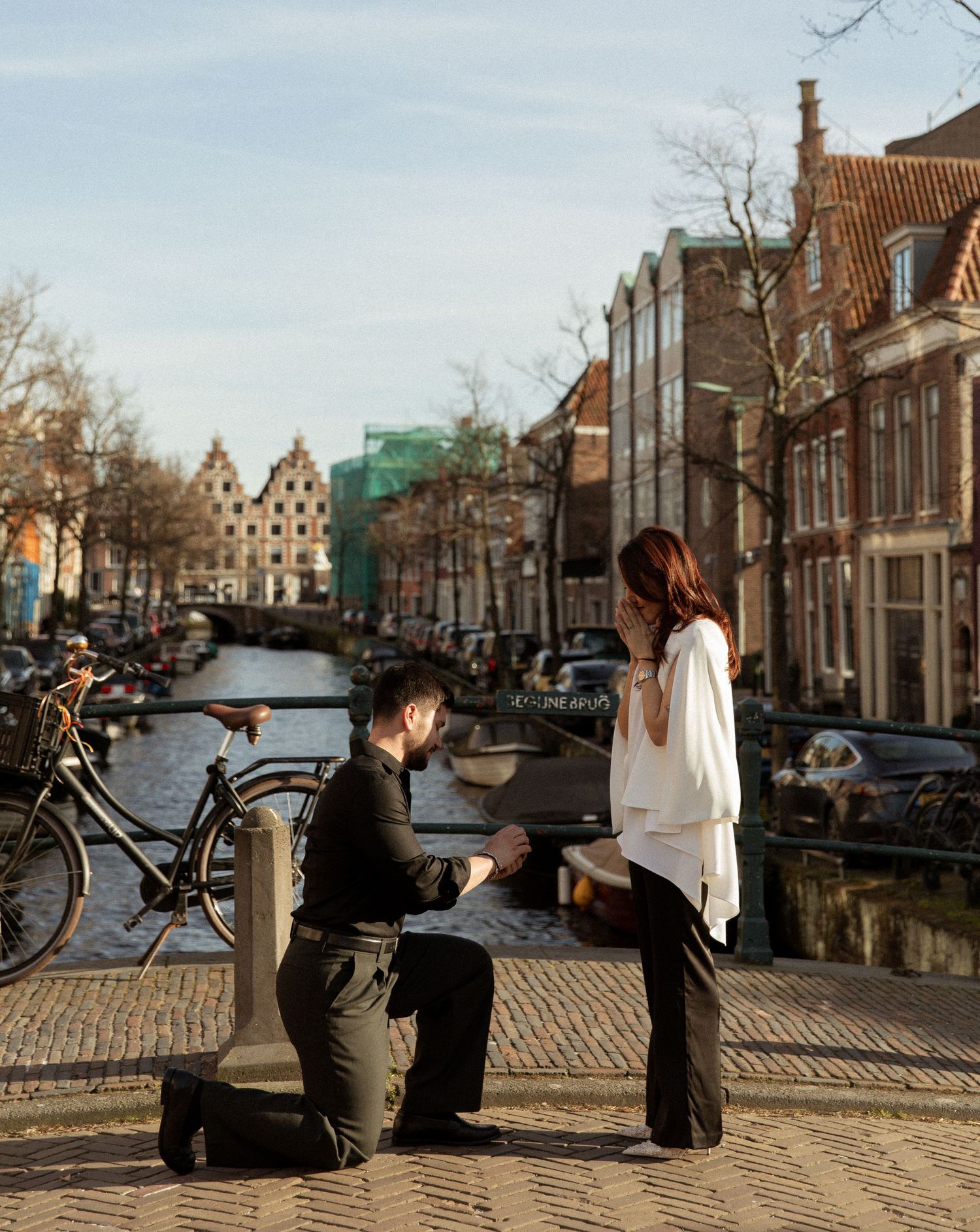 Man proposing to woman on one knee along Amsterdam canal with historic Dutch architecture and bicycles in background.