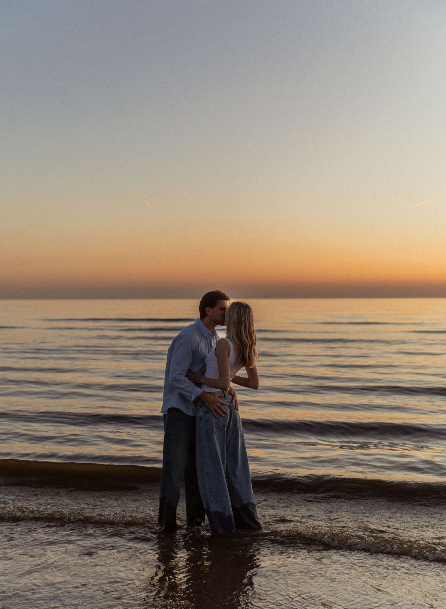 Couple sharing a romantic kiss on the beach at sunset with golden sky and calm ocean waves.