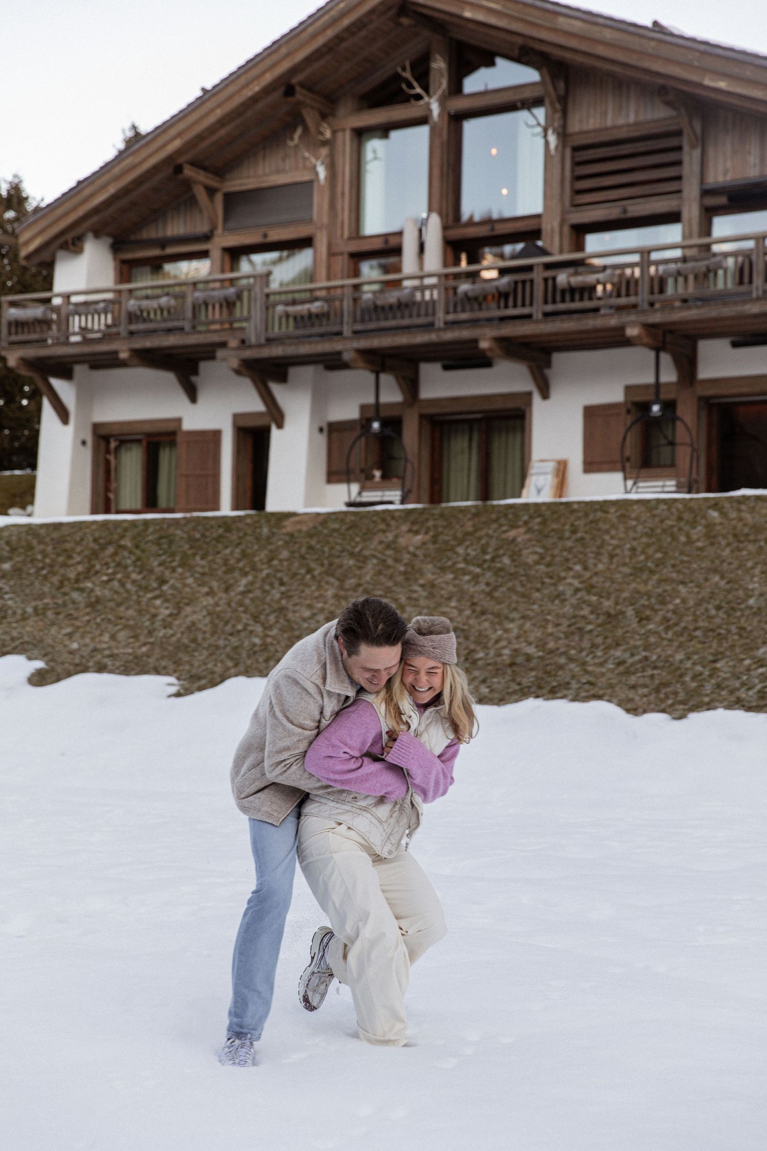 A couple sharing a playful moment in the snow outside a rustic mountain lodge.