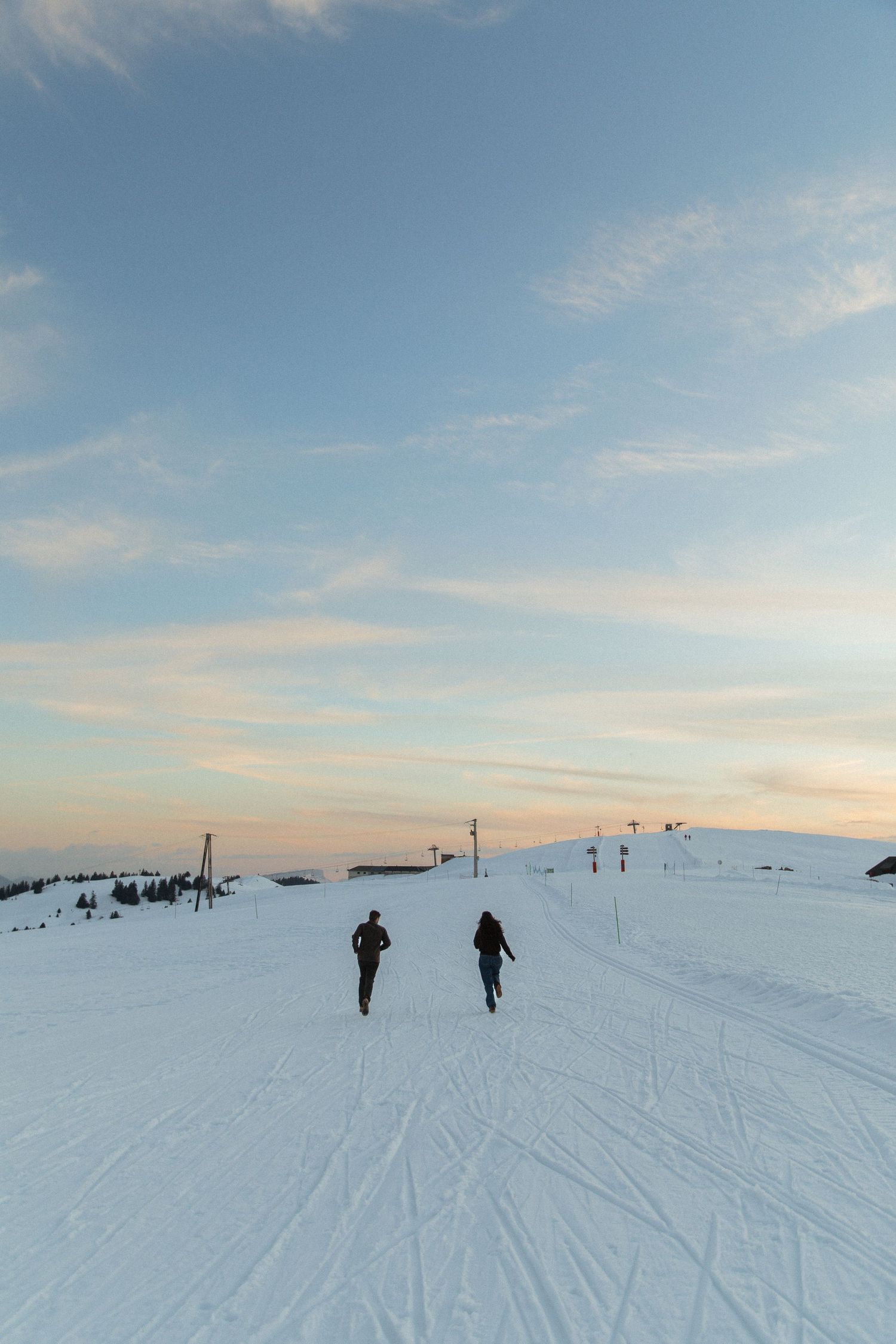 People trek across a vast snowy landscape under pastel winter skies at sunset with ski tracks visible in the snow.