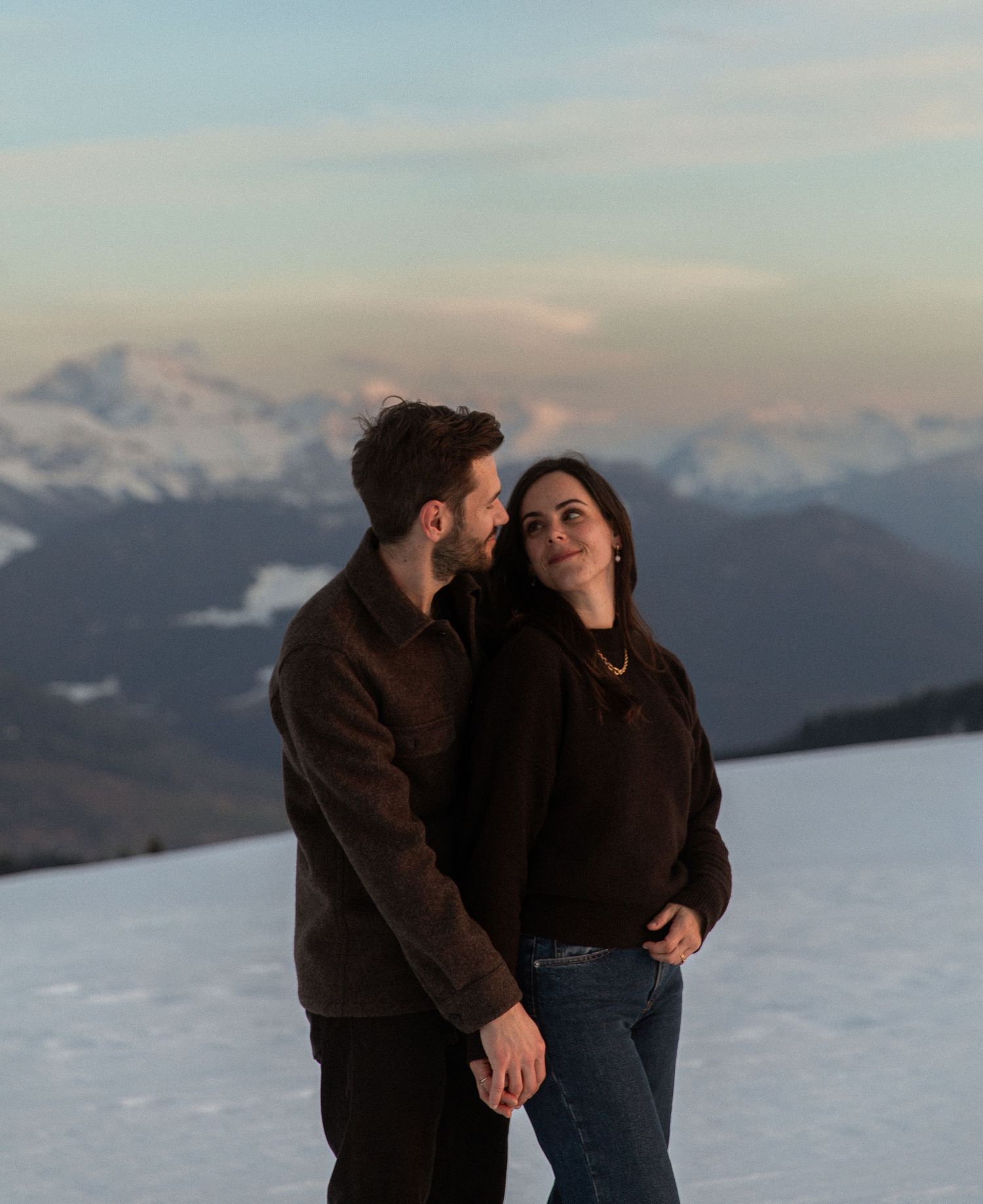 A couple shares an intimate moment against a snowy mountain backdrop at sunset.