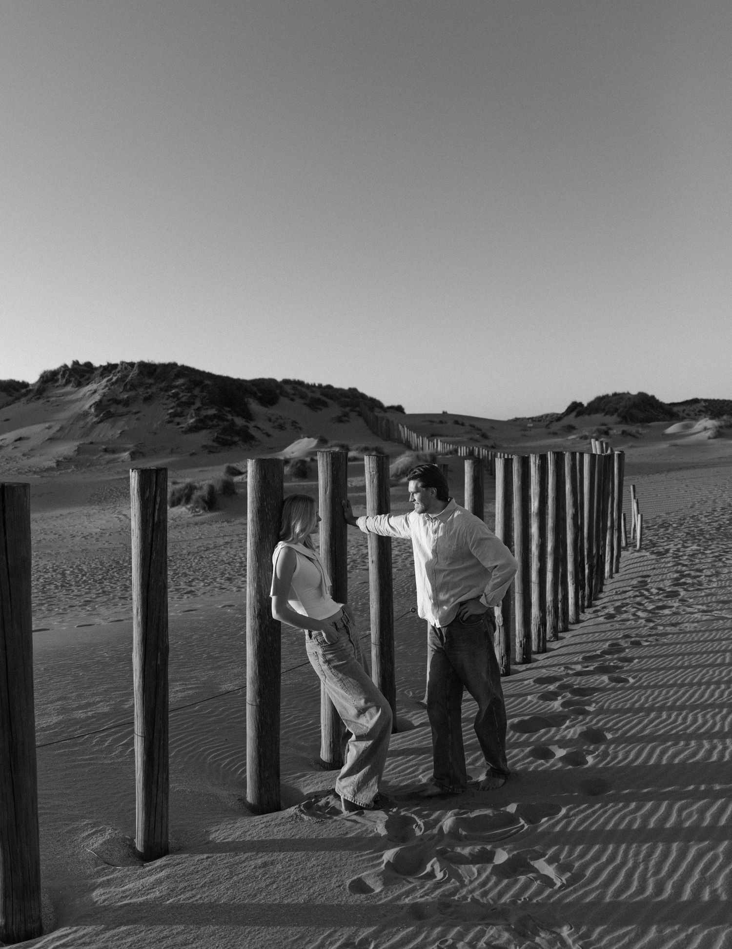 Two people stand among wooden posts in sandy dunes, captured in black and white photography.
