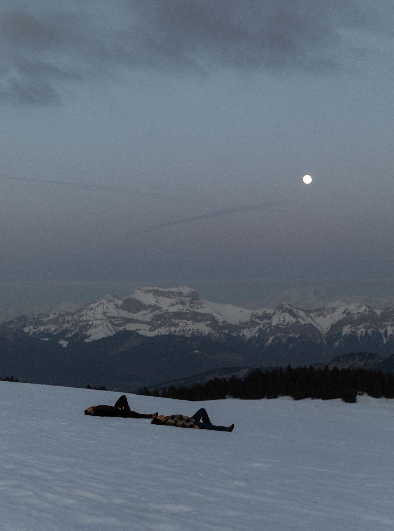 Snow-covered mountain landscape at dusk with silhouetted buildings against a moody gray winter sky.