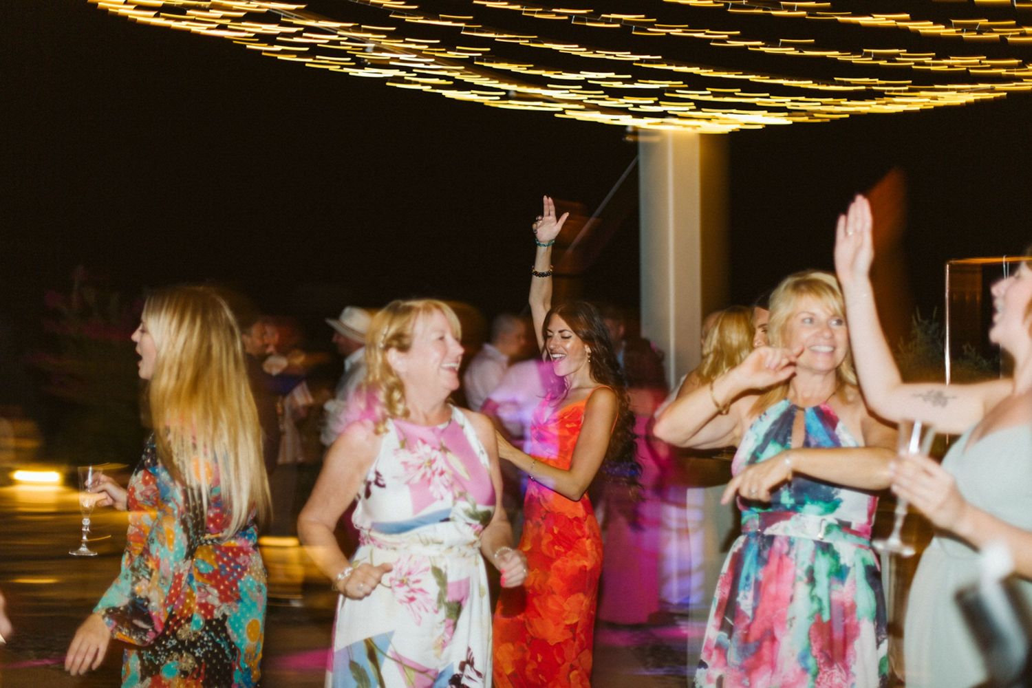 People dancing and celebrating at a wedding party in Santorini with colorful dresses and festive lighting overhead.