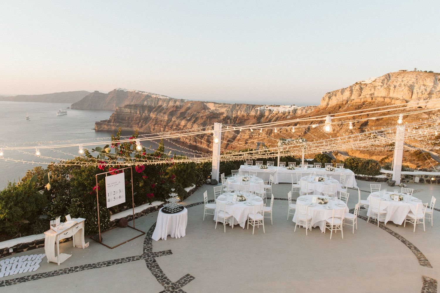 Panoramic view of an elegant outdoor wedding reception setup in Santorini, Greece with white tables overlooking the caldera at sunset.