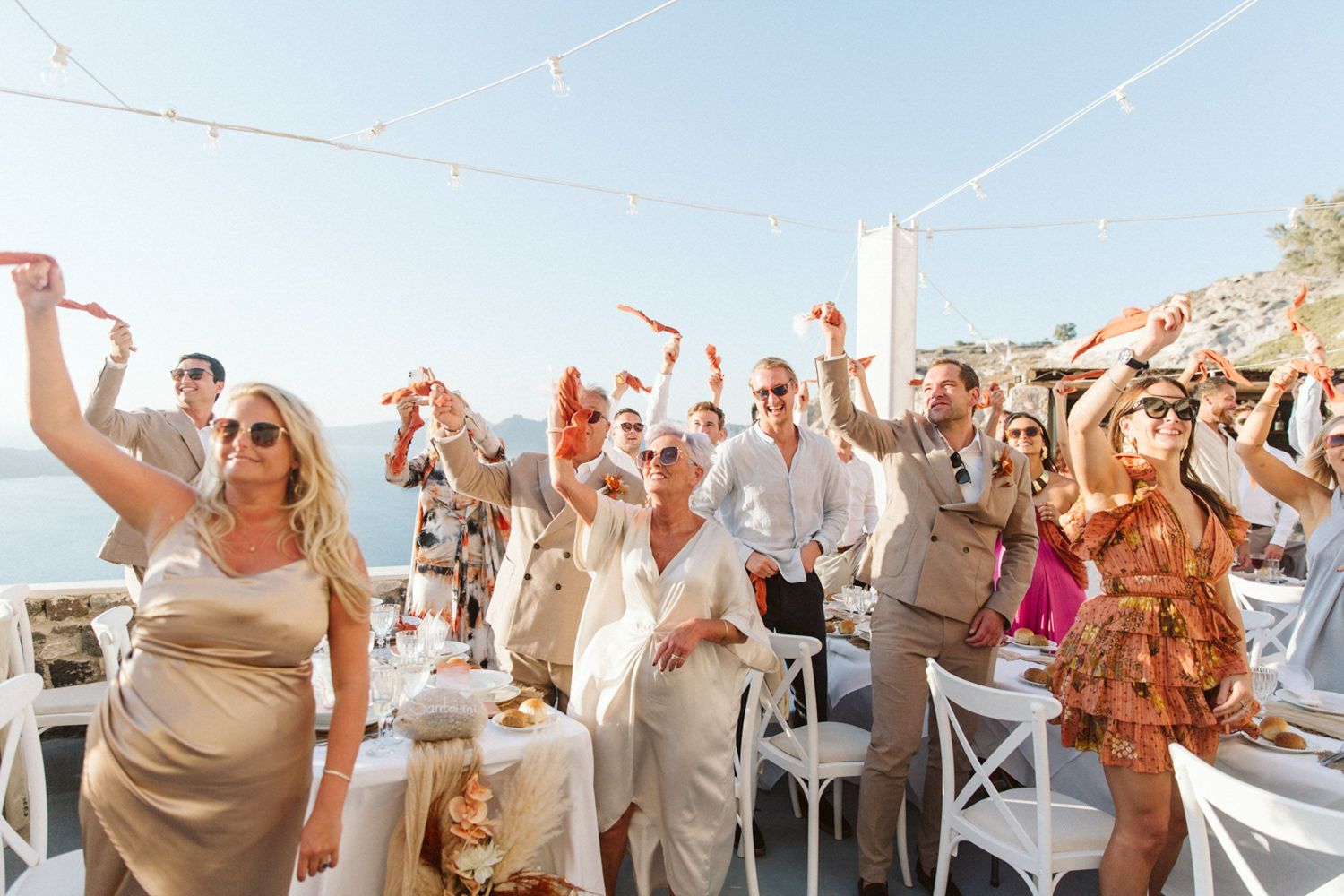 Wedding guests celebrate joyfully at an outdoor reception venue in Santorini overlooking the ocean on a sunny day.