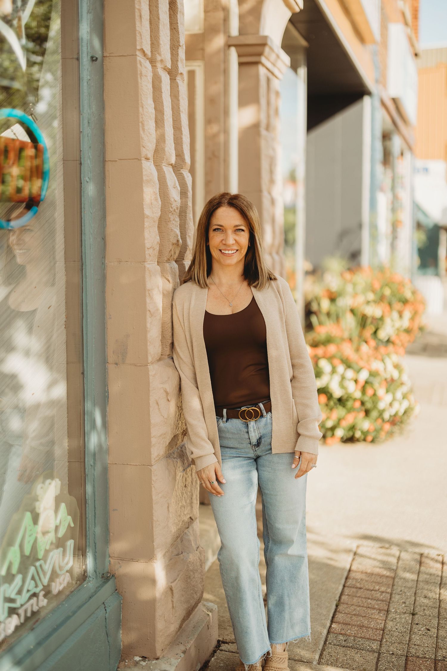 Woman in beige blazer and jeans smiling while leaning against a stone building on a sunny sidewalk.