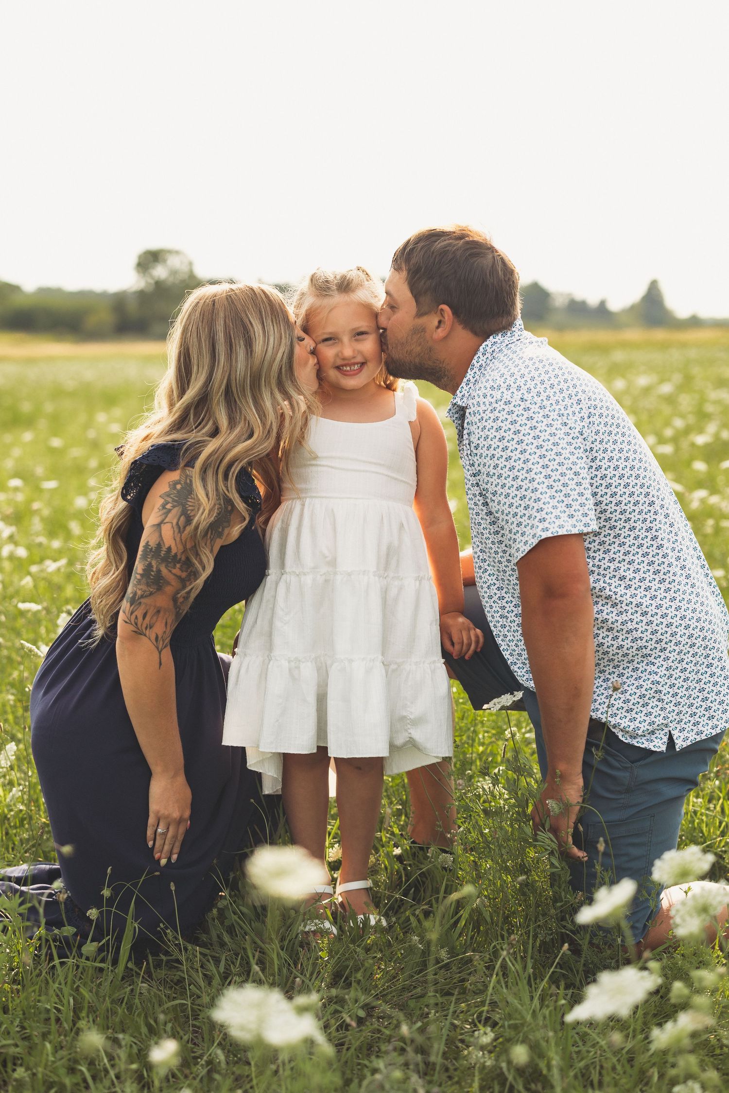 A happy family with parents kissing their young daughter in a sunny flower field, girl in white dress smiling.
