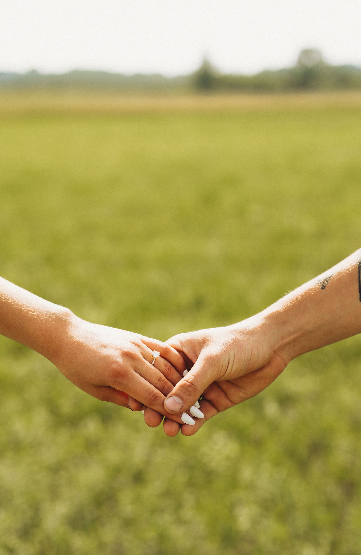 Two people holding hands in a green field, one wearing a ring, symbolizing love and connection outdoors.