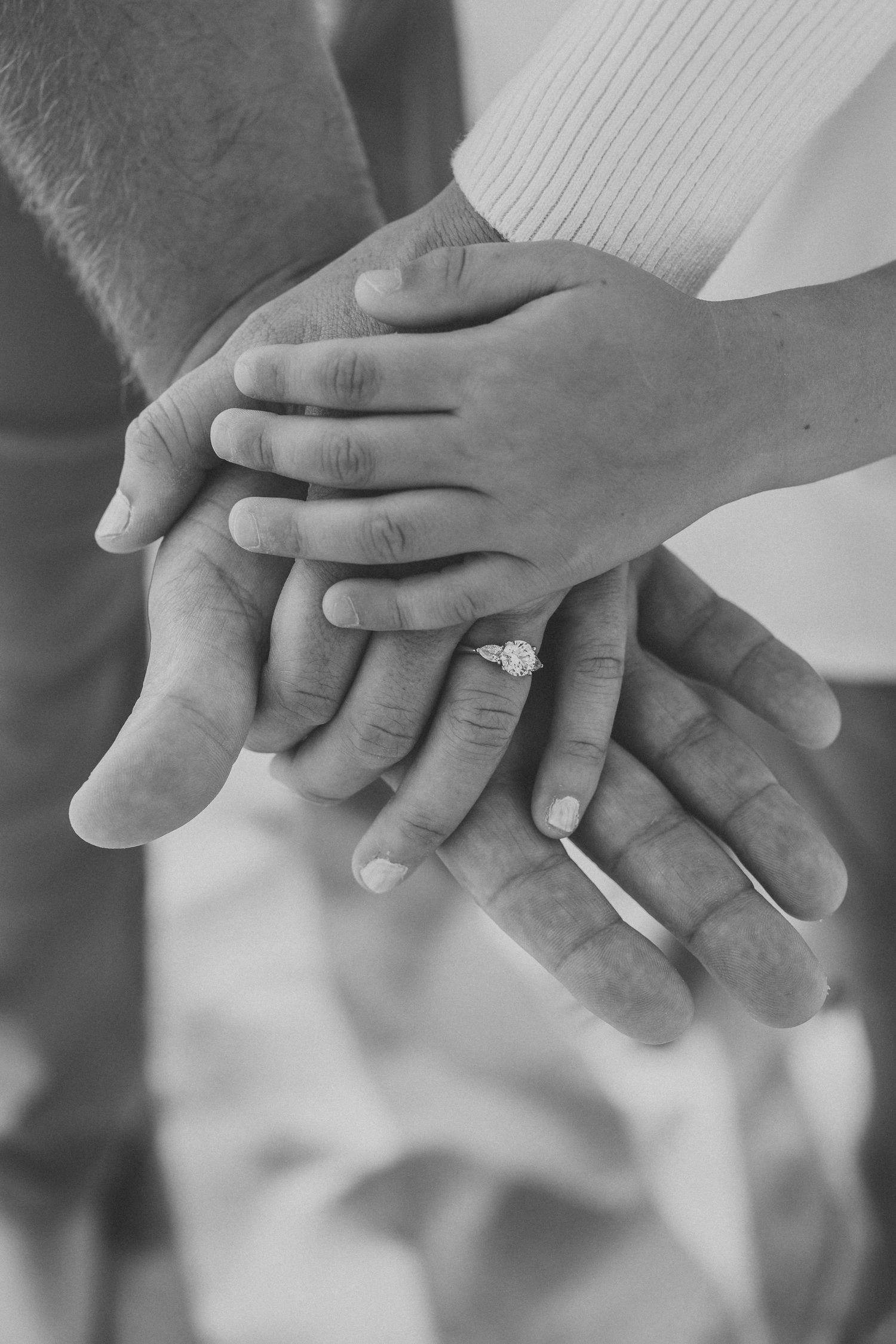 Black and white photo of multiple hands stacked together, with a diamond engagement ring visible on one hand.