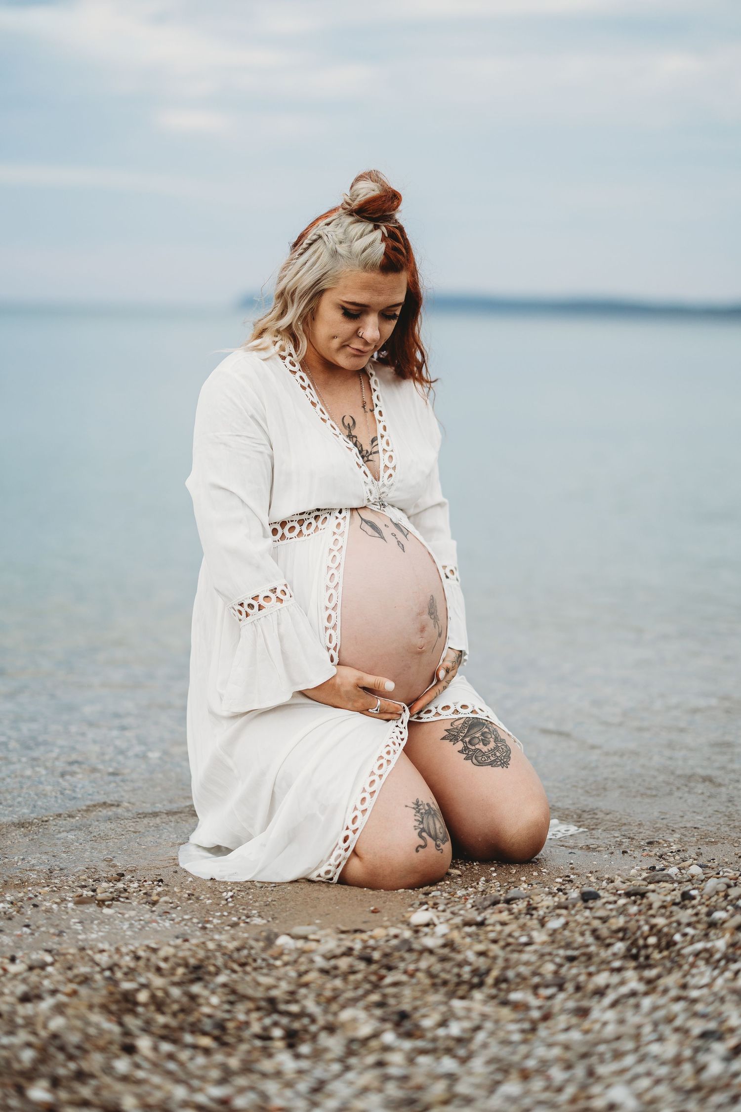 Pregnant woman in white dress kneeling on pebble beach, cradling her bare belly with tattoos visible on her thigh.