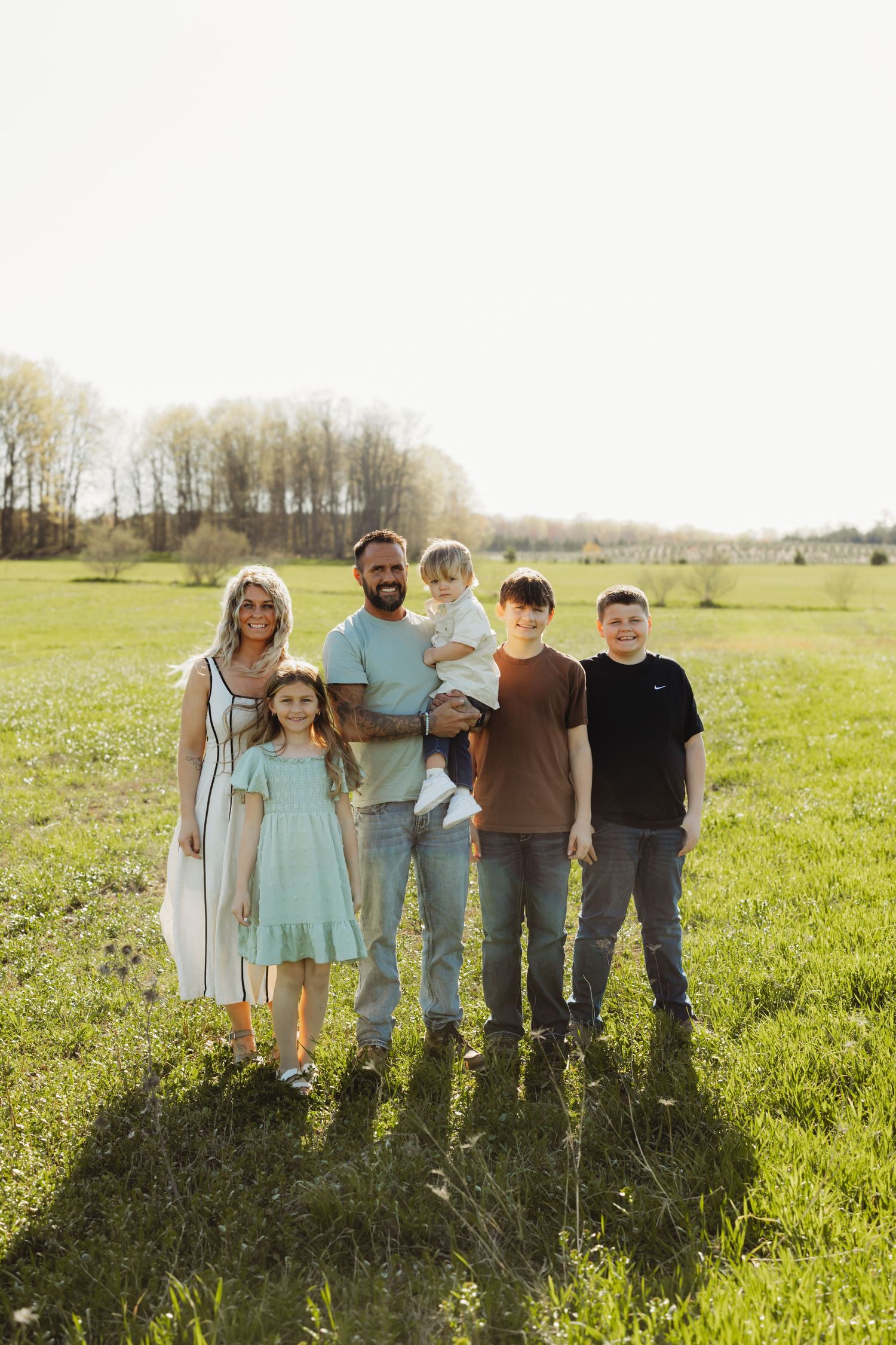Family of six posing together in a sunny green field, with trees in the background on a bright spring day.